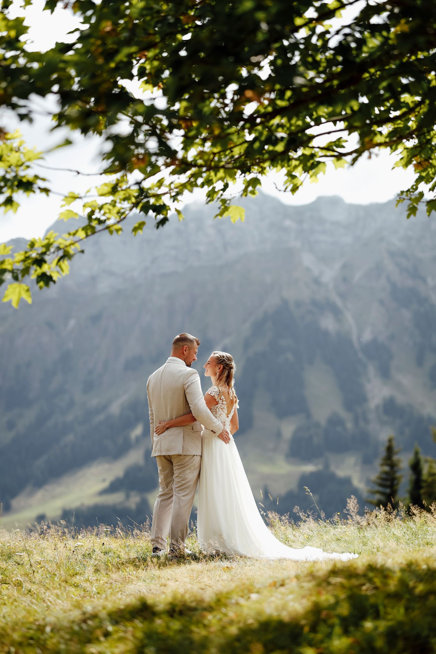 A bride and groom stand together under a tree with breathtaking mountain scenery behind them.