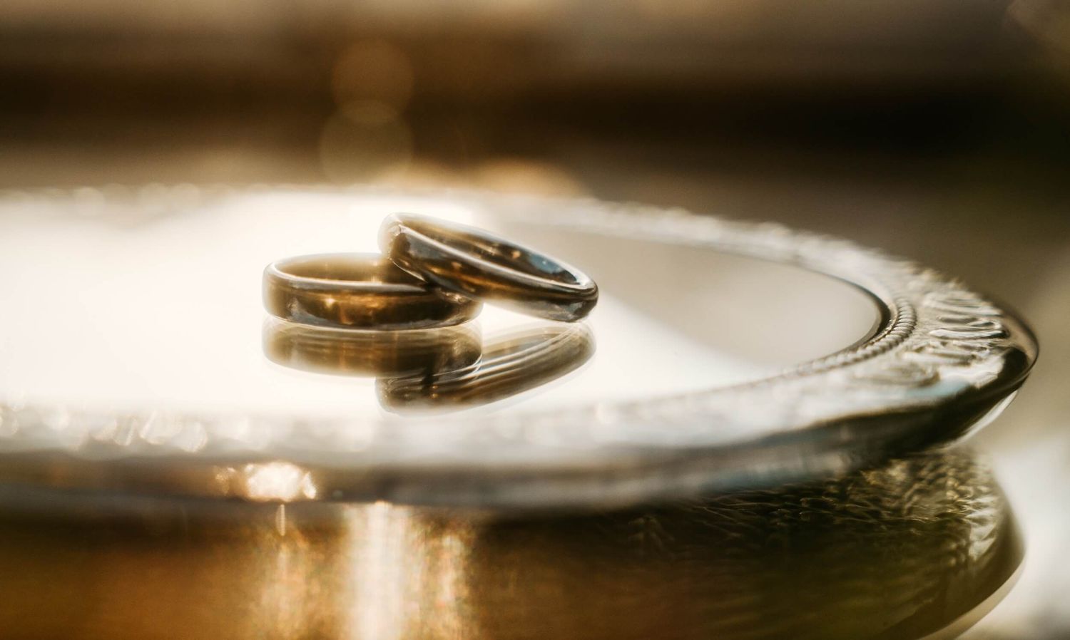 Two gold and silver wedding bands resting on an ornate silver plate in warm golden light.