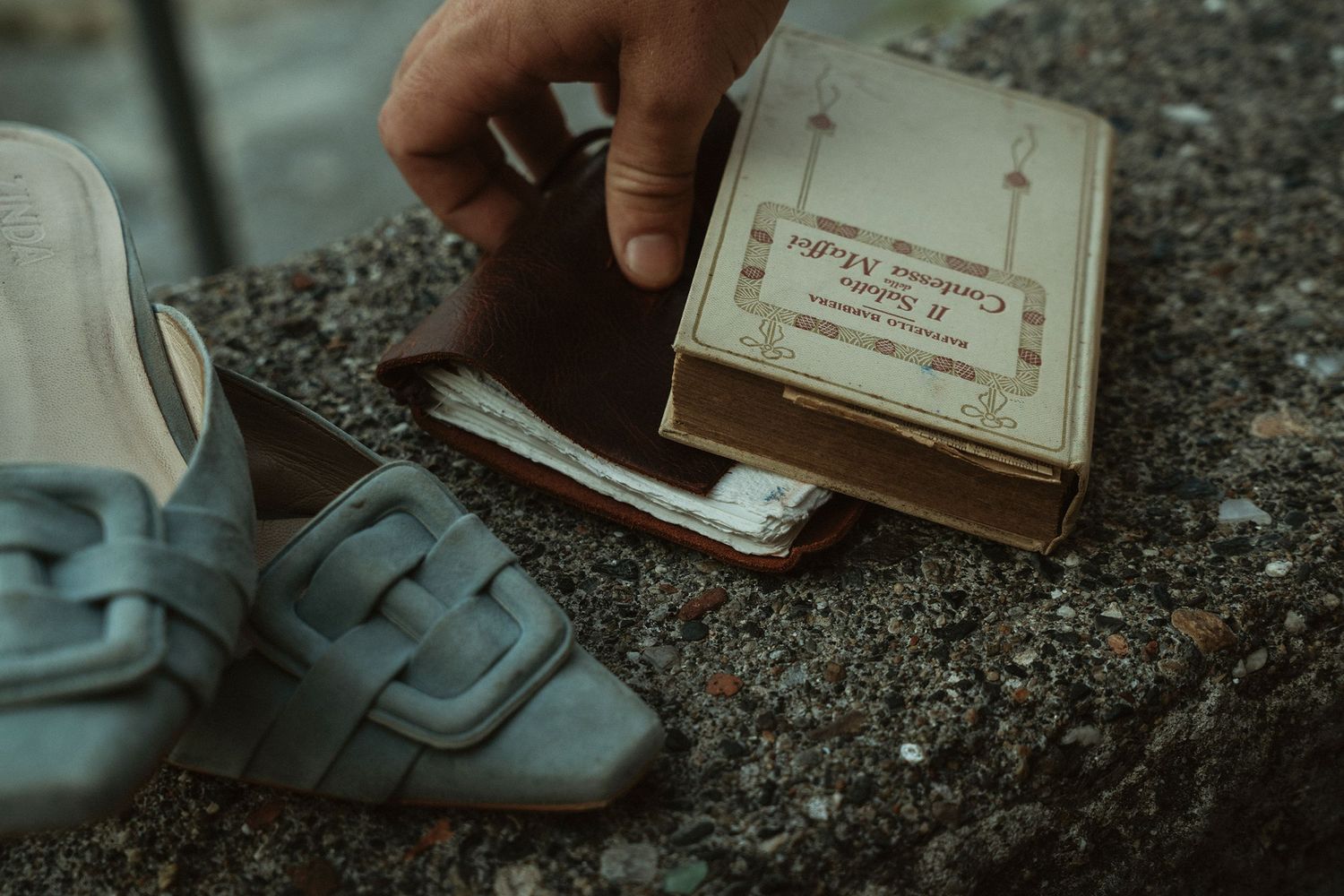 Hand holding a vintage box next to leather shoes and cash on a stone surface.