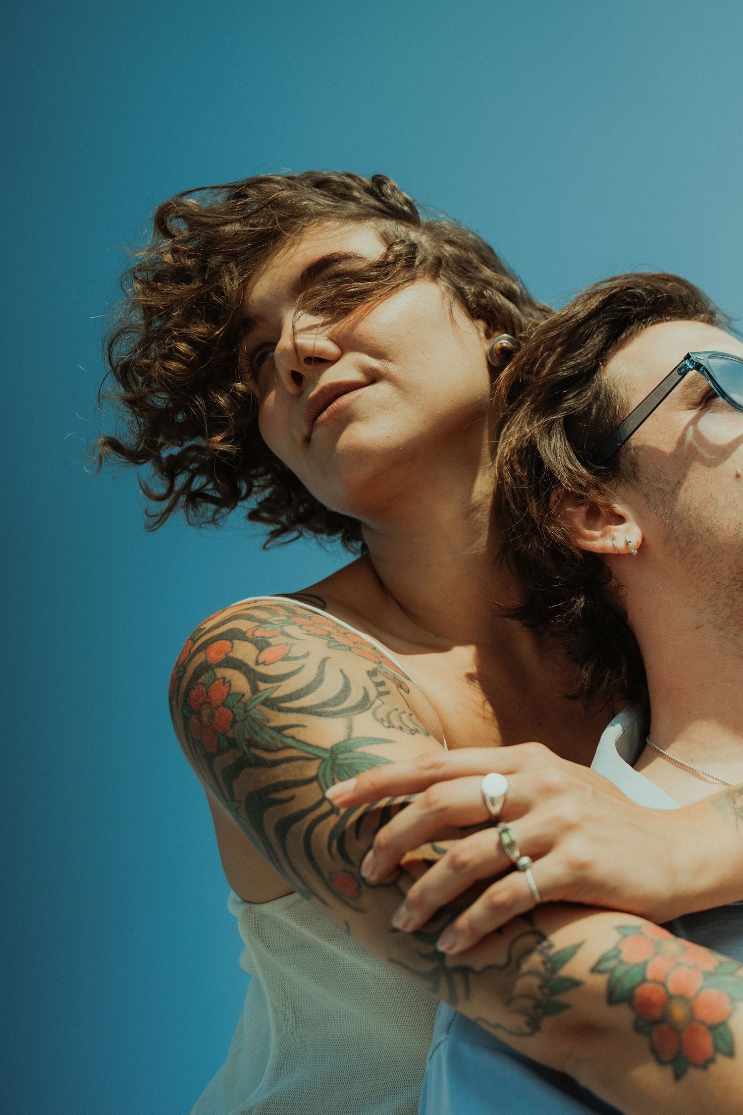 Tattooed couple embracing against vibrant blue sky, woman in white dress leaning back on mans shoulder.