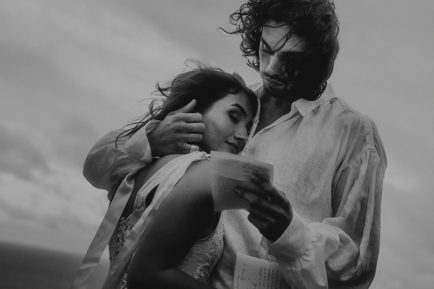 Black and white photo of a romantic couple embracing outdoors, woman reading a letter as man holds her close.