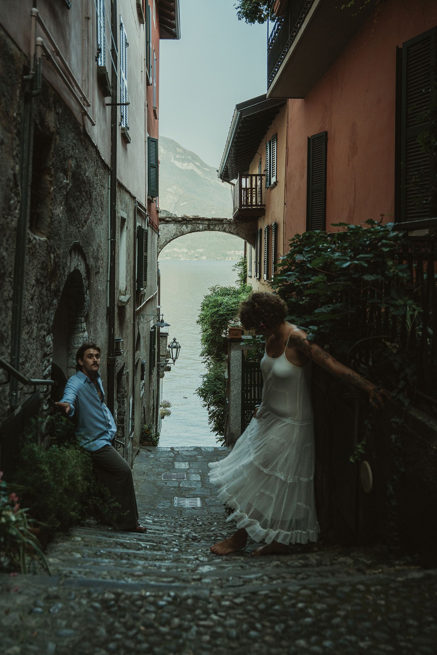 Couple in a charming Italian cobblestone alley with colorful buildings and lake view.