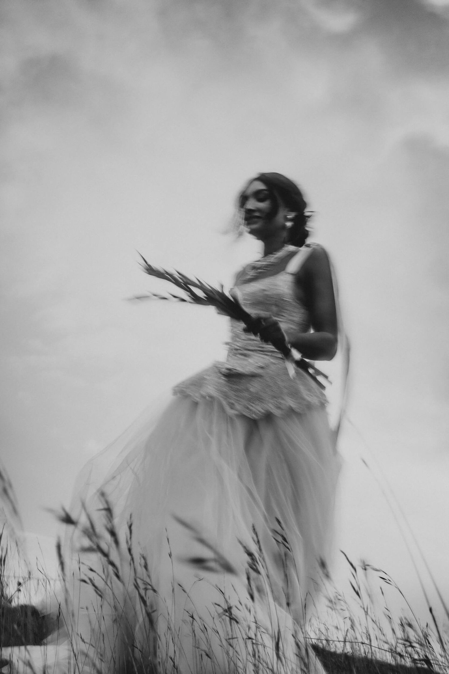 Black and white blurred photo of a bride in a flowing gown holding wildflowers in a field.