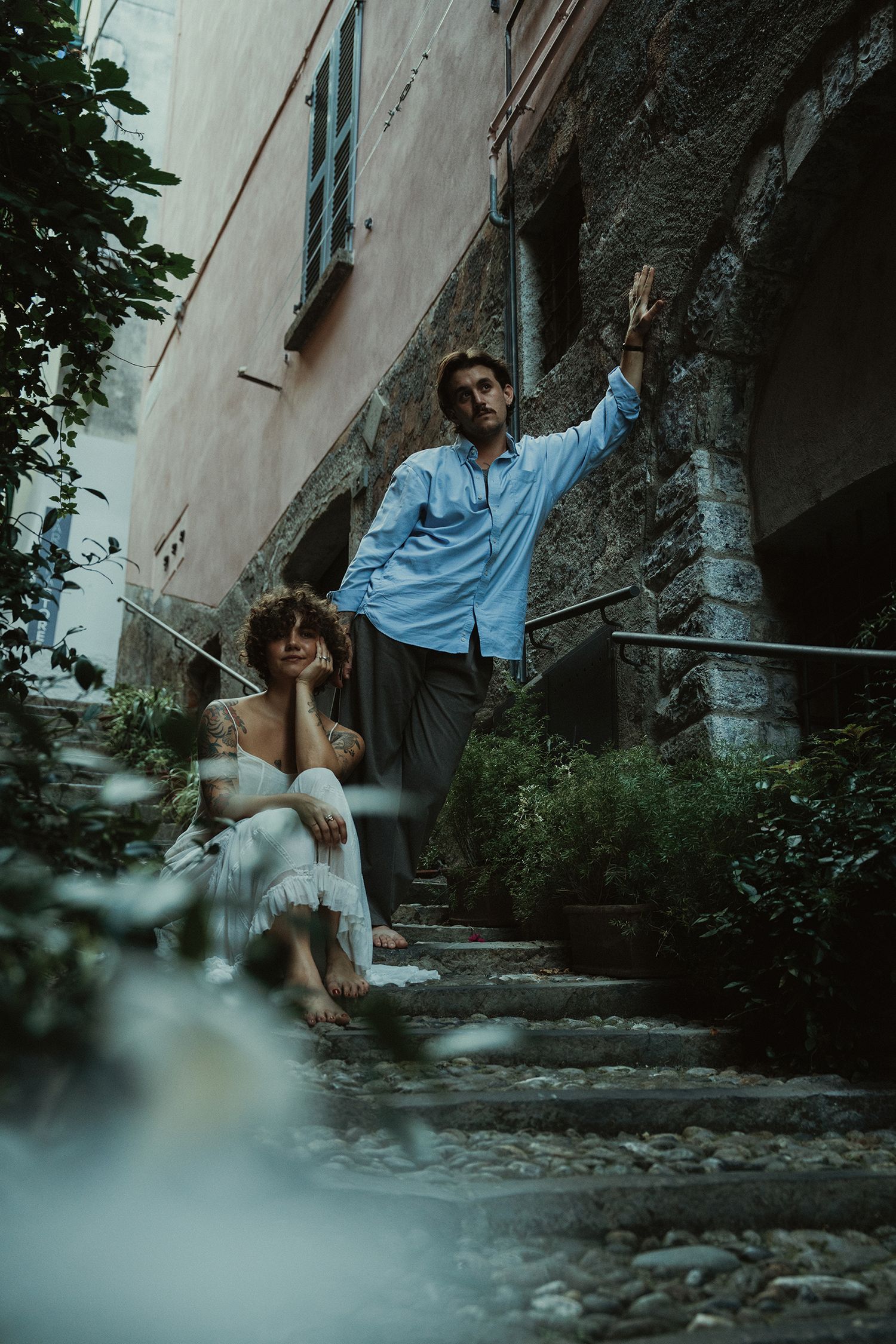 Couple posing on rustic stone steps surrounded by lush greenery in an Italian village.