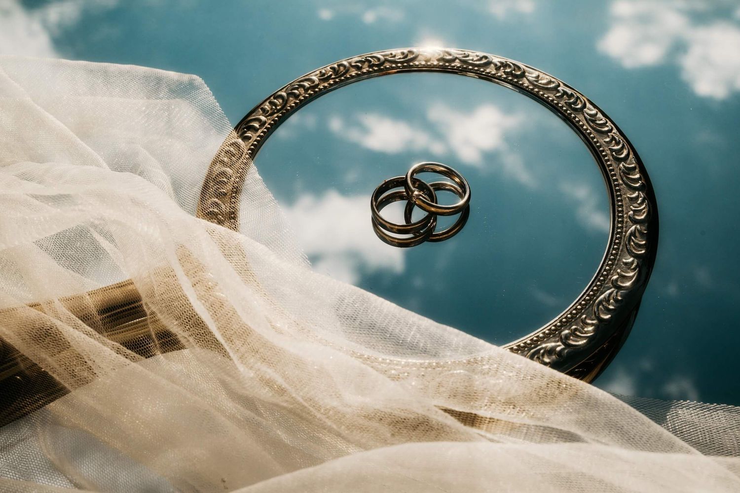 Two gold wedding rings rest inside an ornate circular mirror with a flowing veil and cloudy sky reflection.
