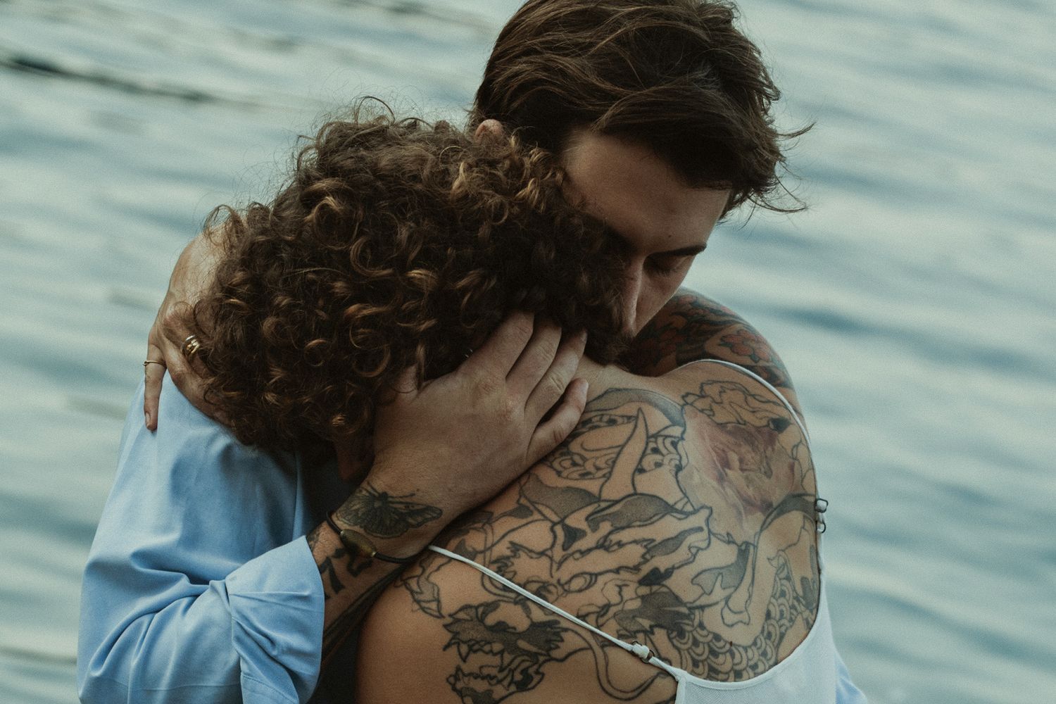 Couple sharing intimate embrace by water, man with back tattoos hugging woman in blue shirt.
