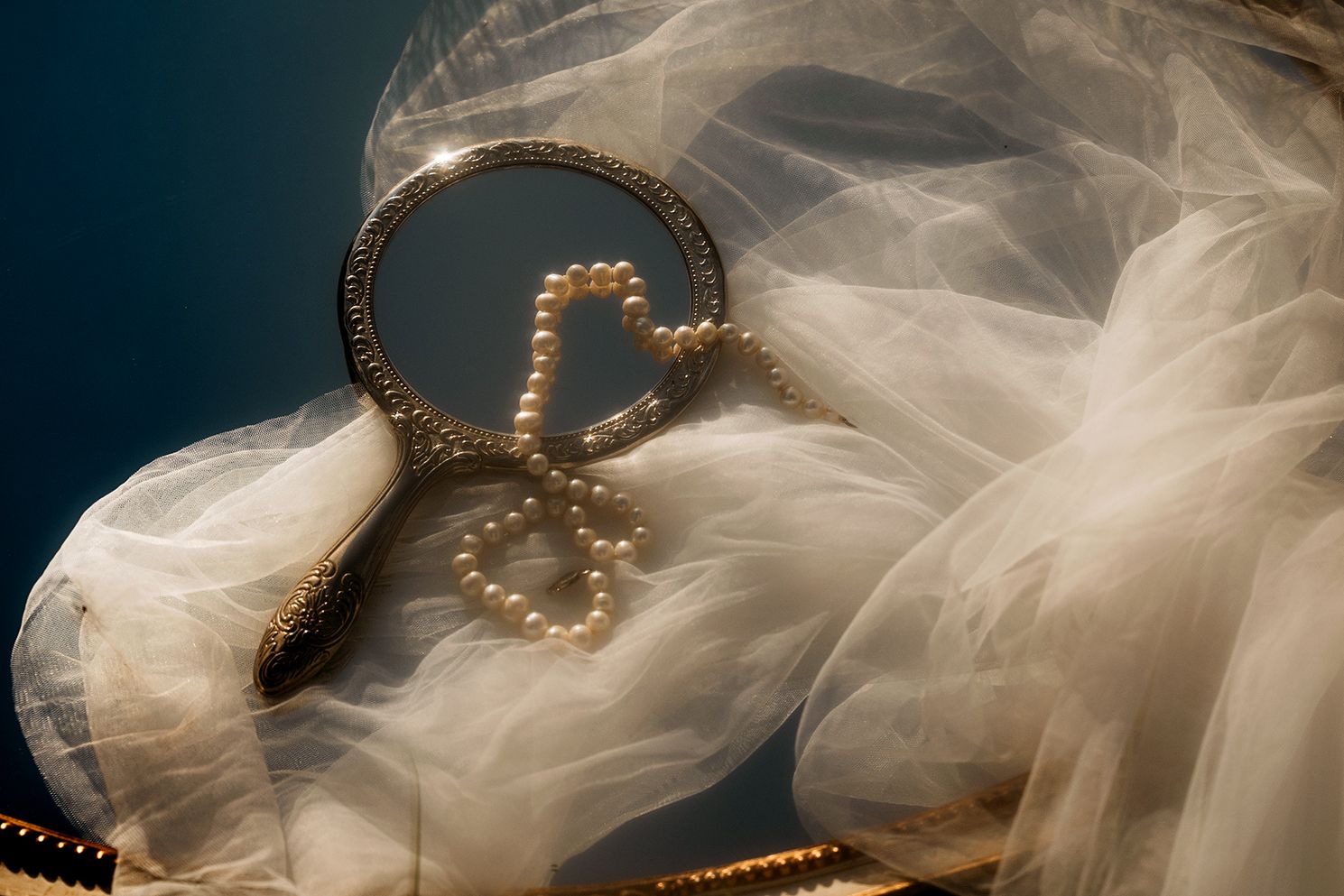 A vintage hand mirror adorned with pearl hearts rests on white tulle fabric, perfect for a bridal setting.