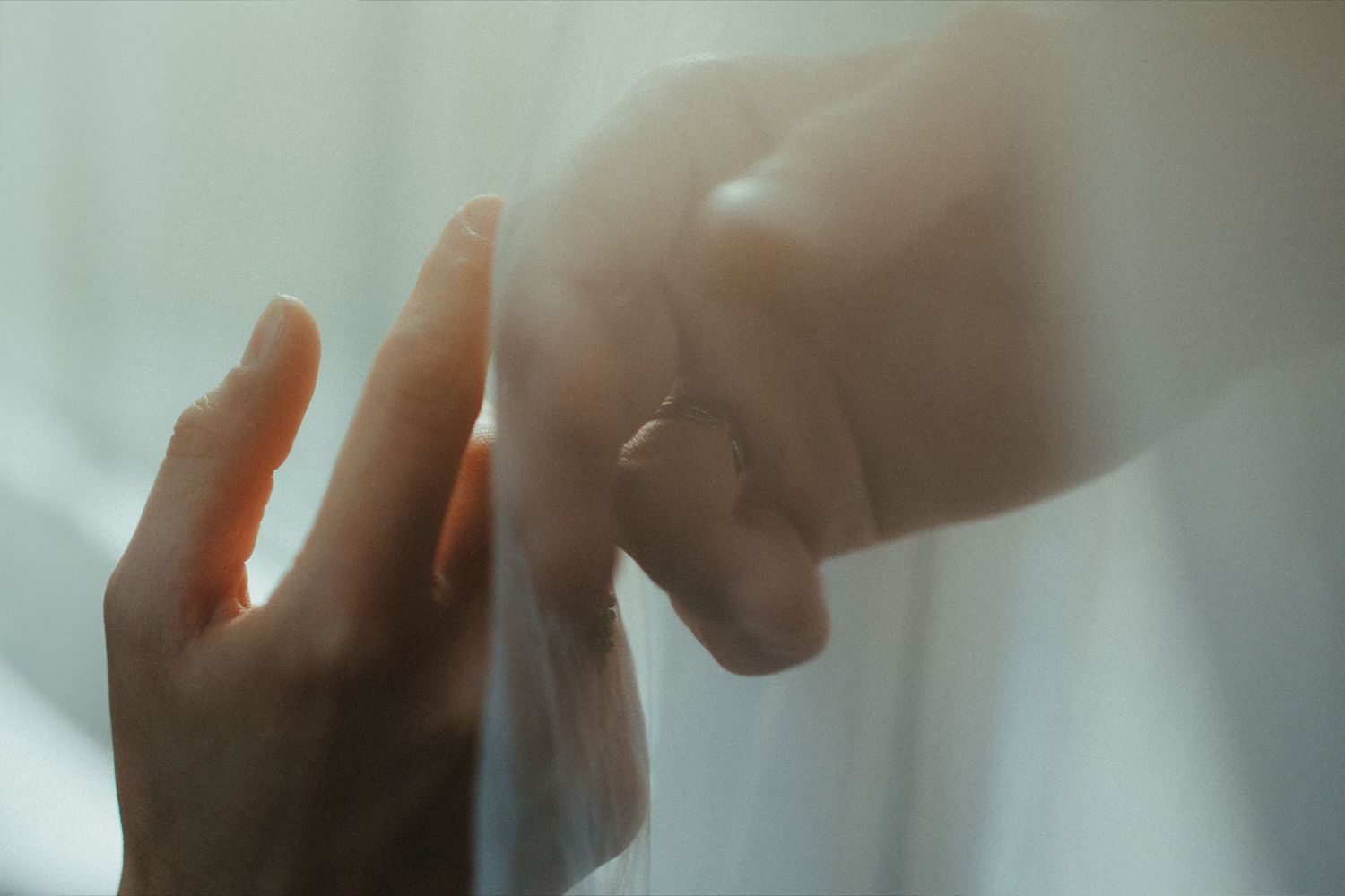 Close-up of two hands gently touching through sheer fabric, one wearing a ring, in soft ethereal light.