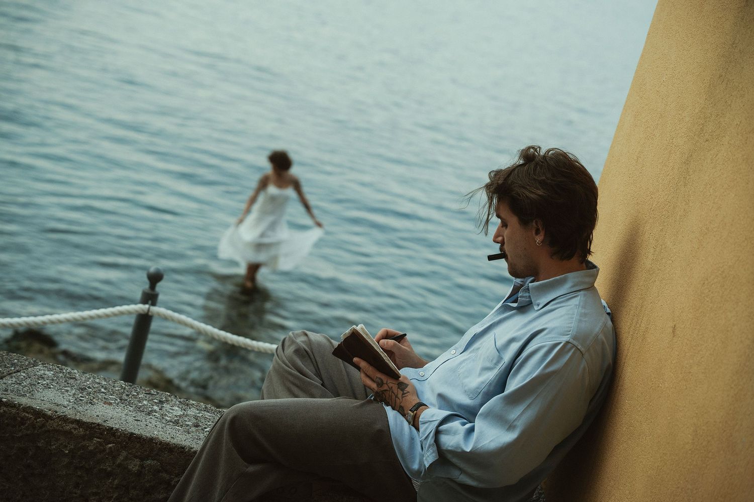 Man reading a book on stone steps by the water while a woman in white dress walks toward the sea.