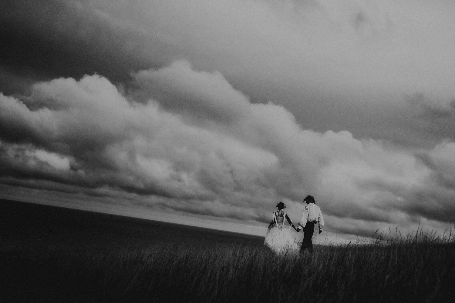 Black and white silhouette of an elopement couple walking through a wheat field under dramatic stormy clouds.