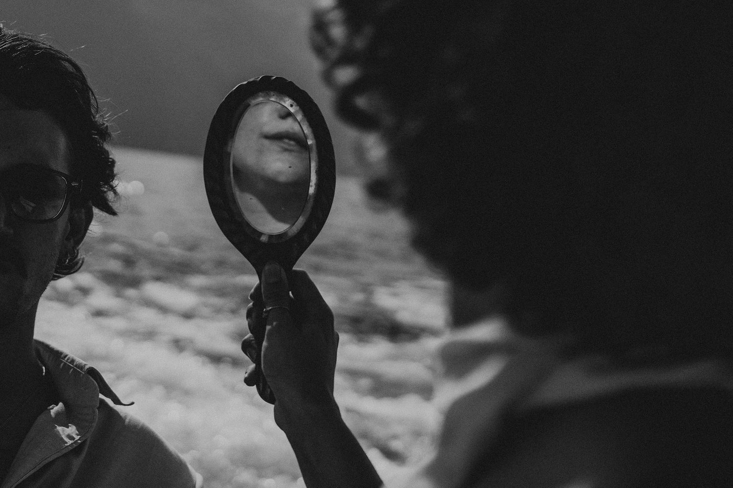 Black and white photo of a woman gazing into a handheld mirror outdoors.