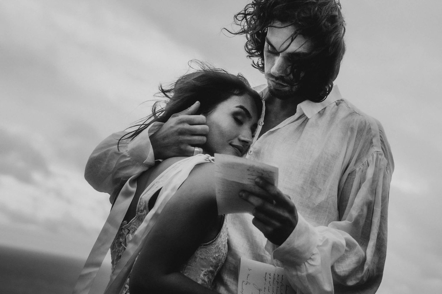 Black and white romantic photo of couple embracing and reading vows outdoors under a cloudy sky.