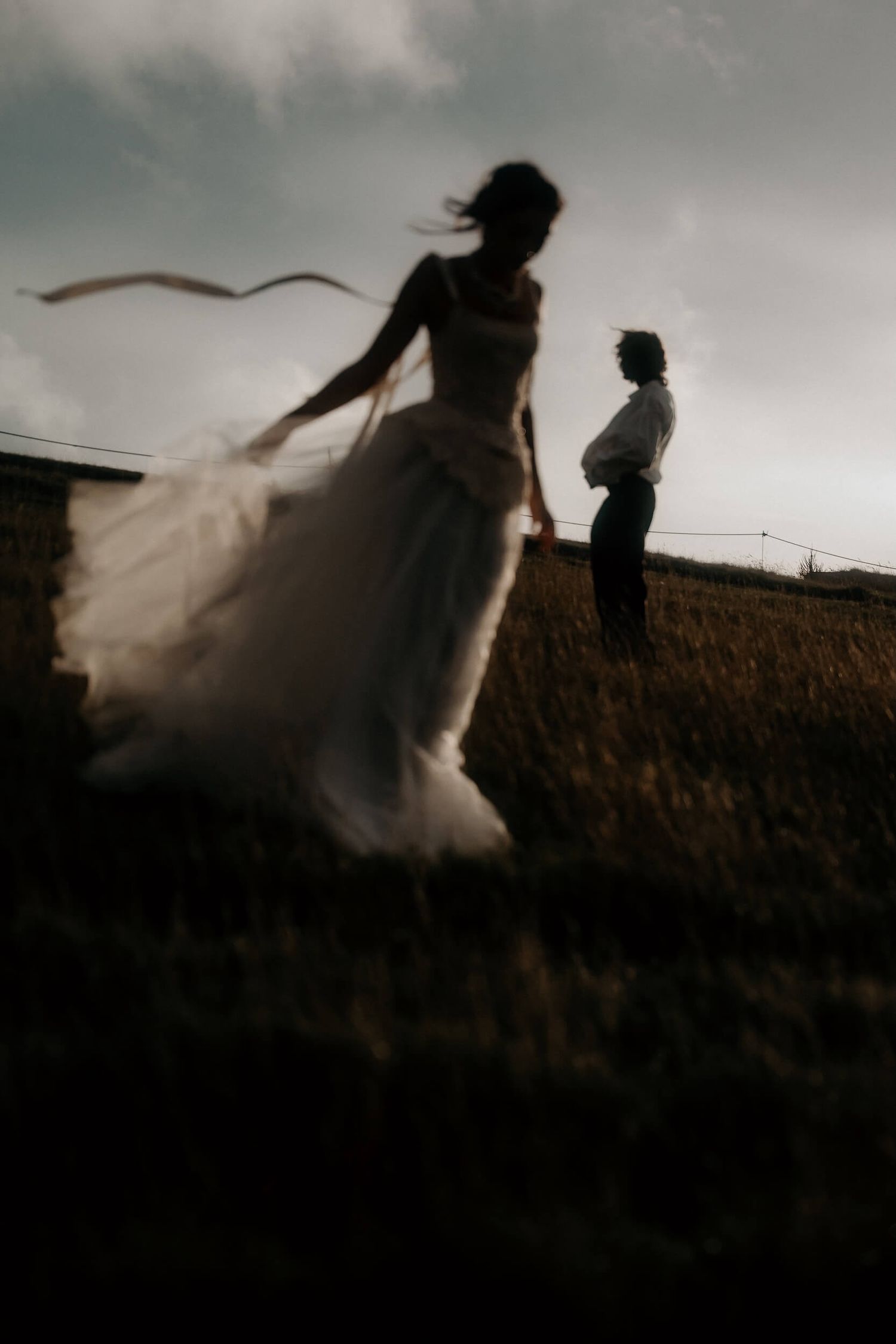 Blurred silhouette of bride in tulle gown and groom standing in dark field at sunset.