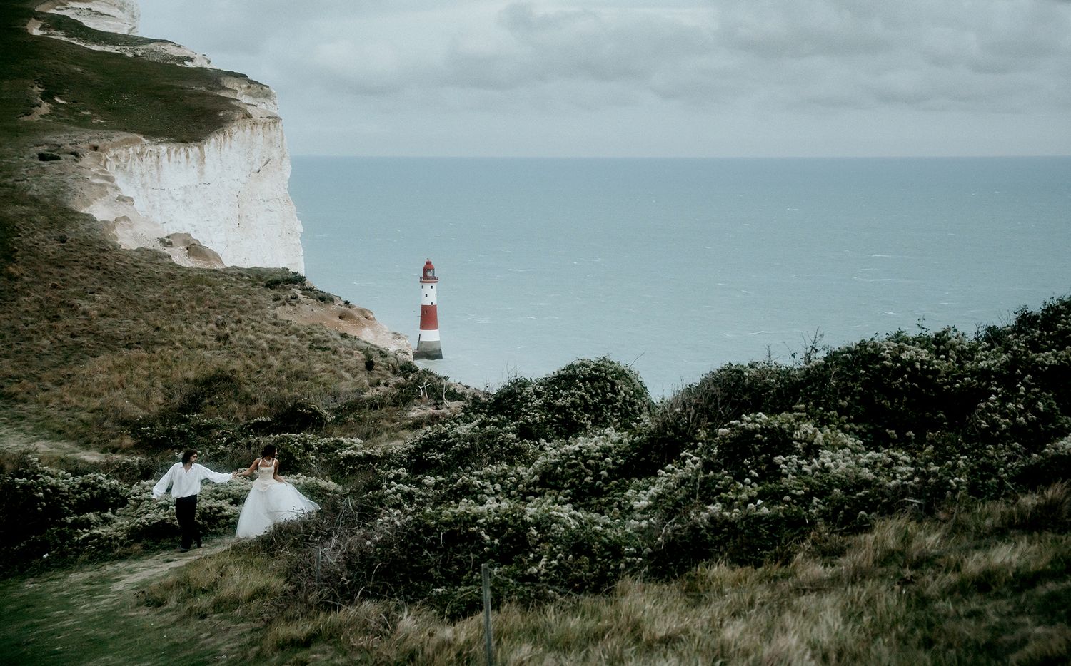 Couple running through green cliffs near Beachy Head lighthouse on cloudy day.