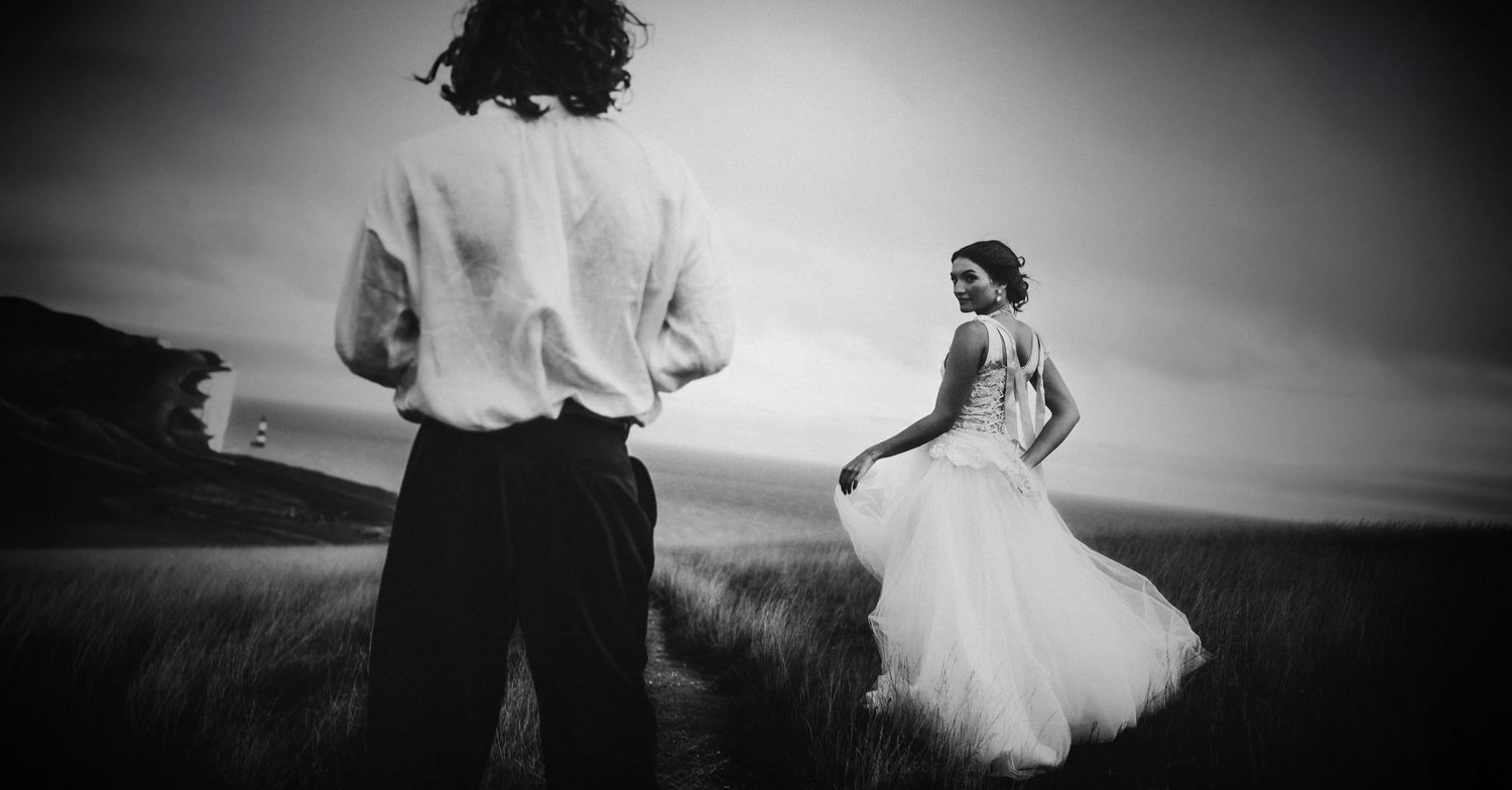 Black and white wedding photo of bride in flowing gown and groom standing in open field at dusk.