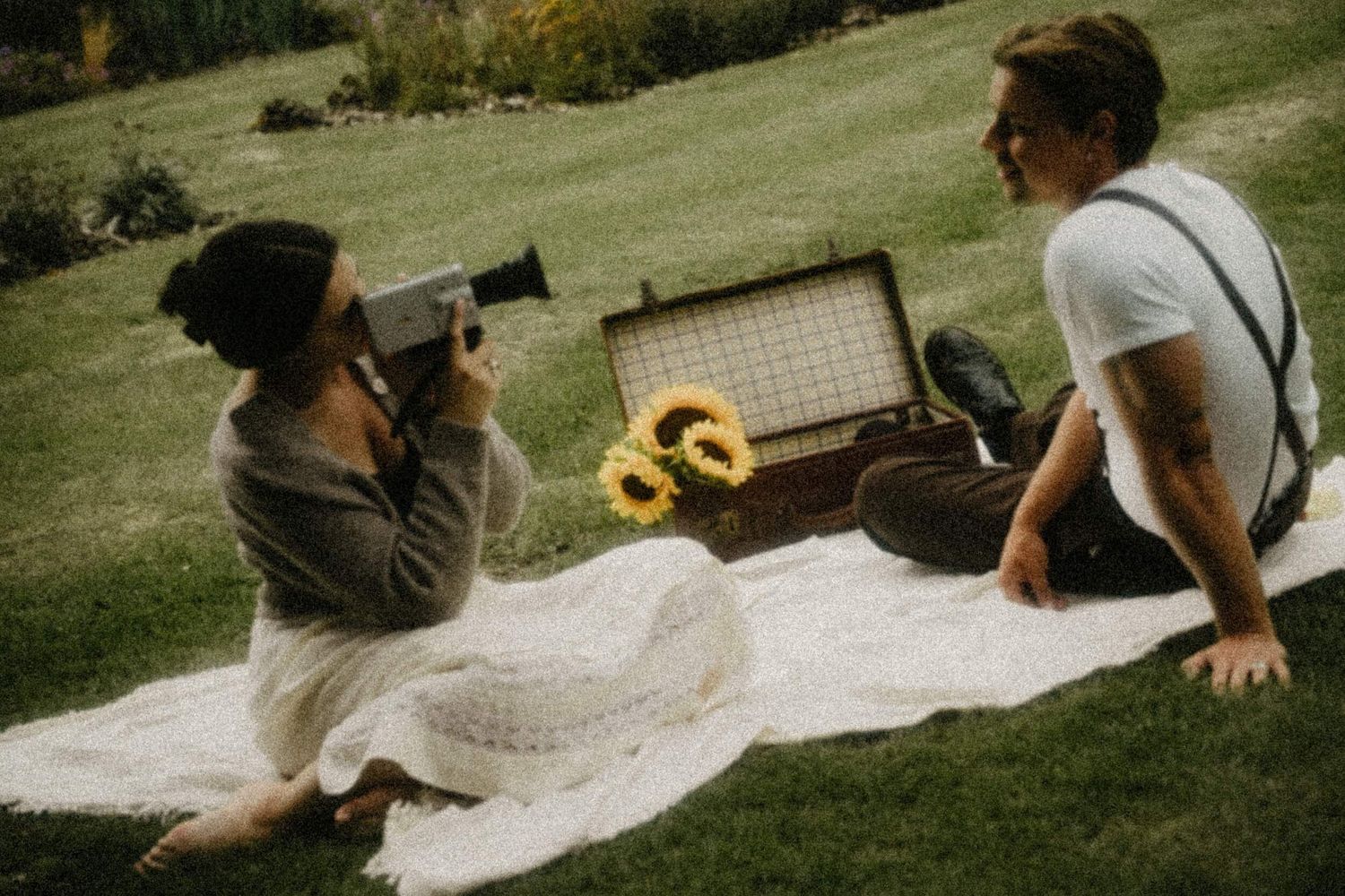 Couple enjoying a vintage picnic on a blanket with sunflowers and a retro radio, woman filming with an old super 8 camera.