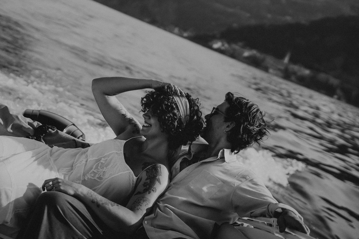 Black and white photo of two people lying close together on a speedboat, with water spray and mountains visible in background.