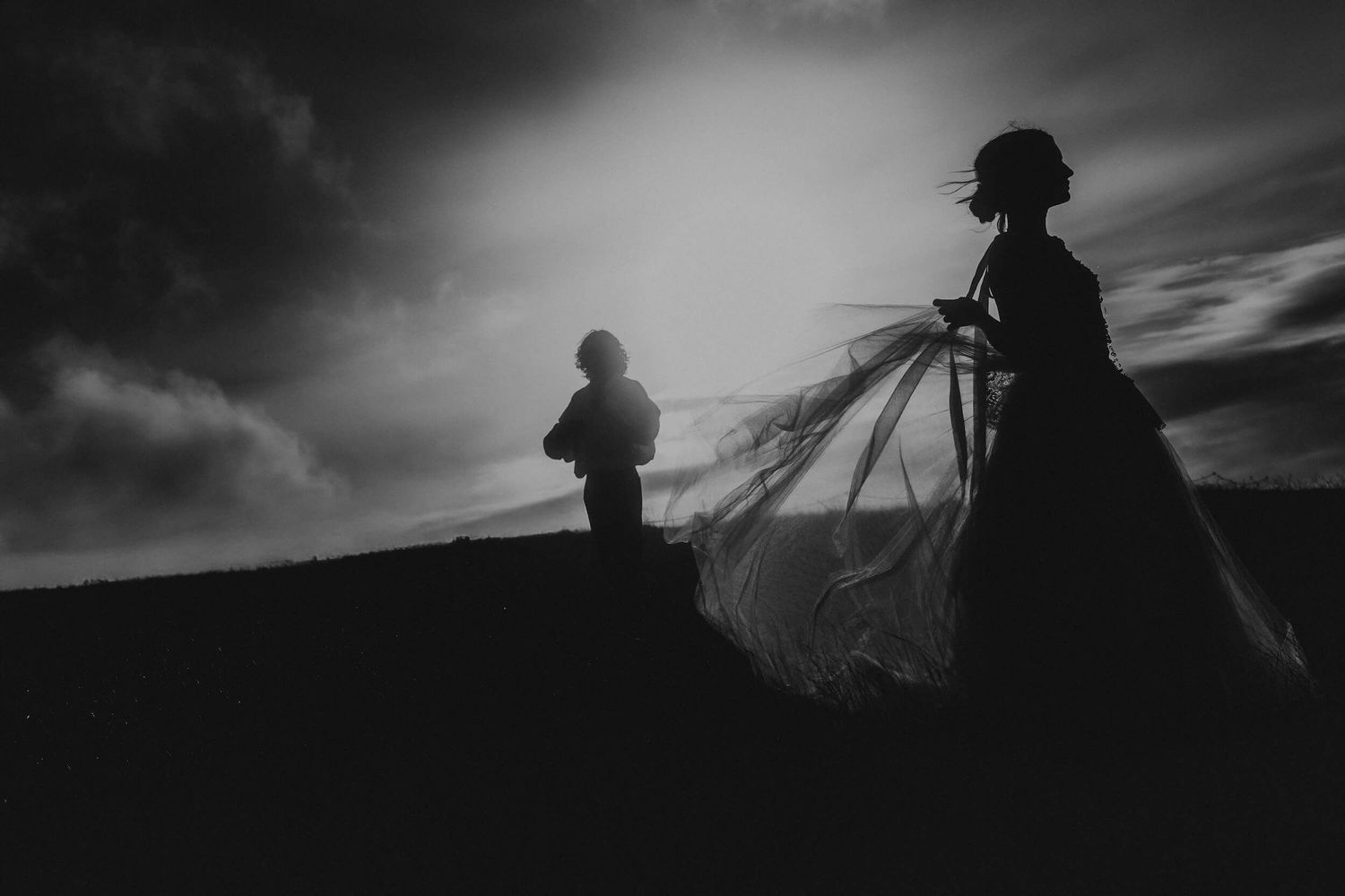 Black and white silhouette of bride and groom on hilltop with dramatic cloudy sky.