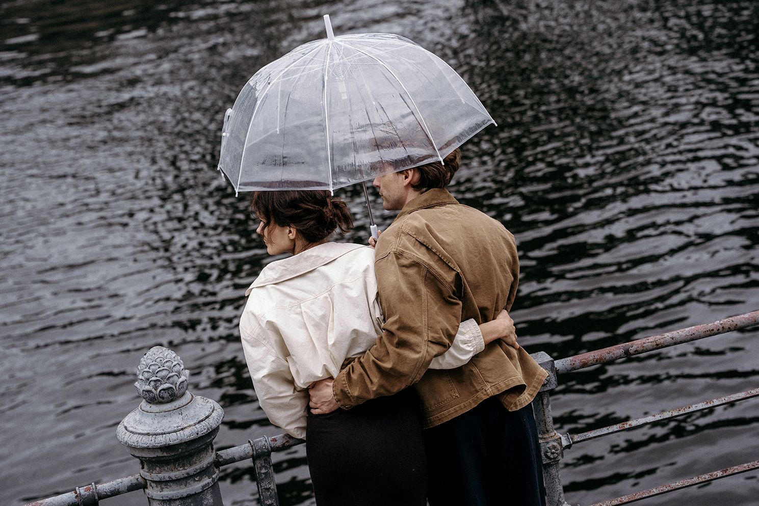 Romantic couple sharing clear umbrella embracing by dark river on rainy day.