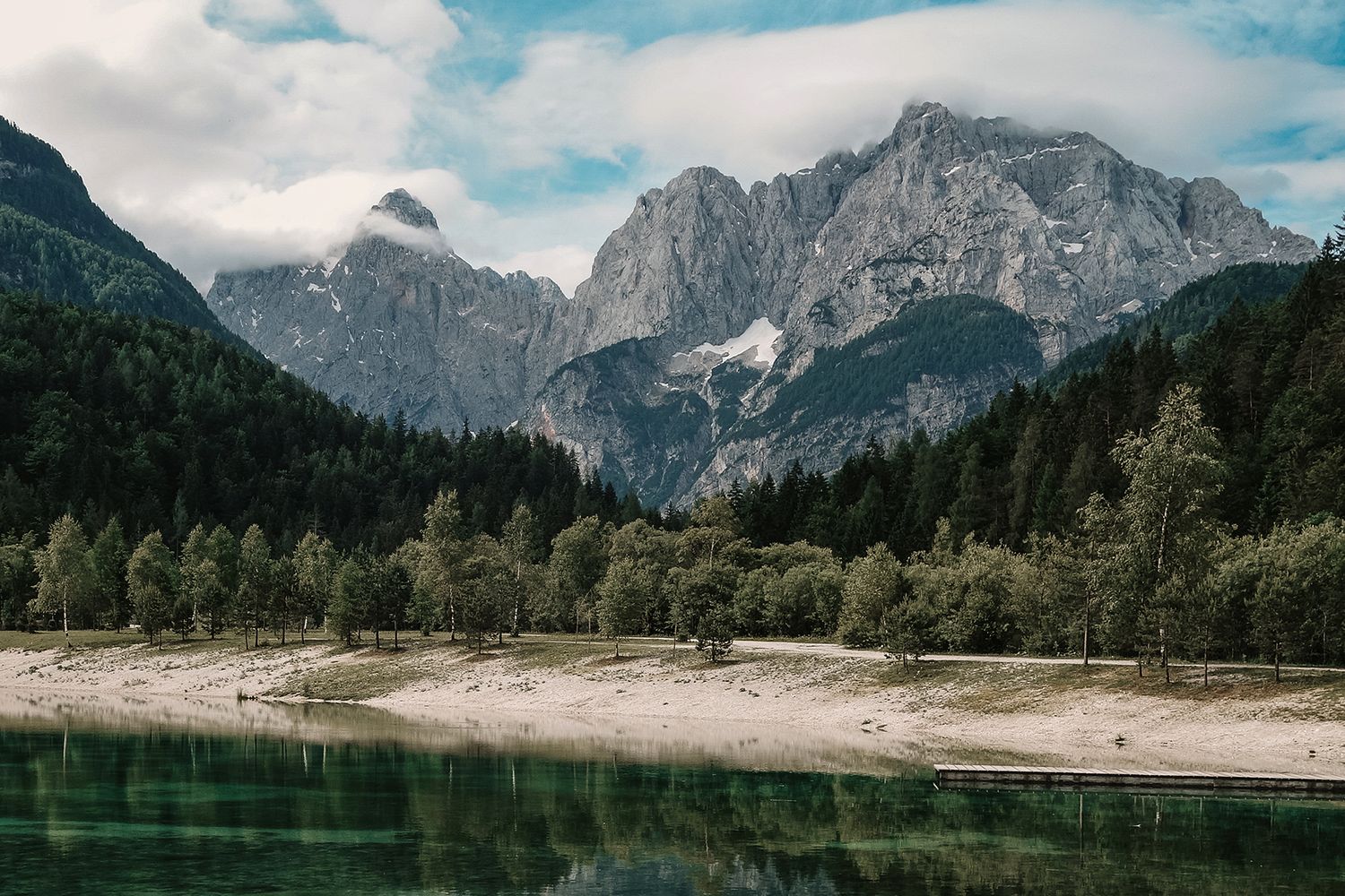 Tranquil mountain lake with turquoise water, sandy shore, and dramatic rocky peaks beyond.