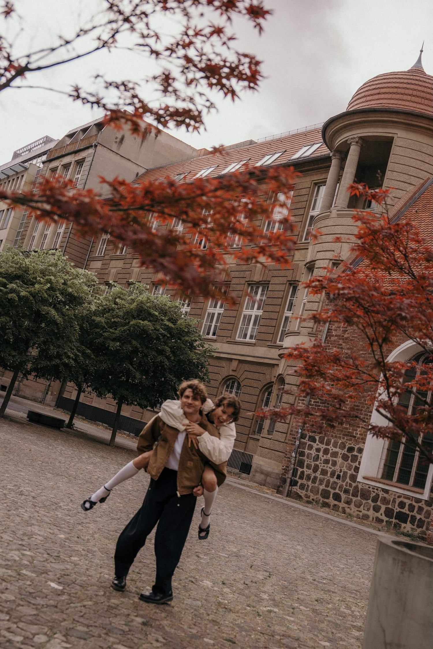 A couple playfully piggyback riding in a cobblestone courtyard surrounded by autumn red trees.