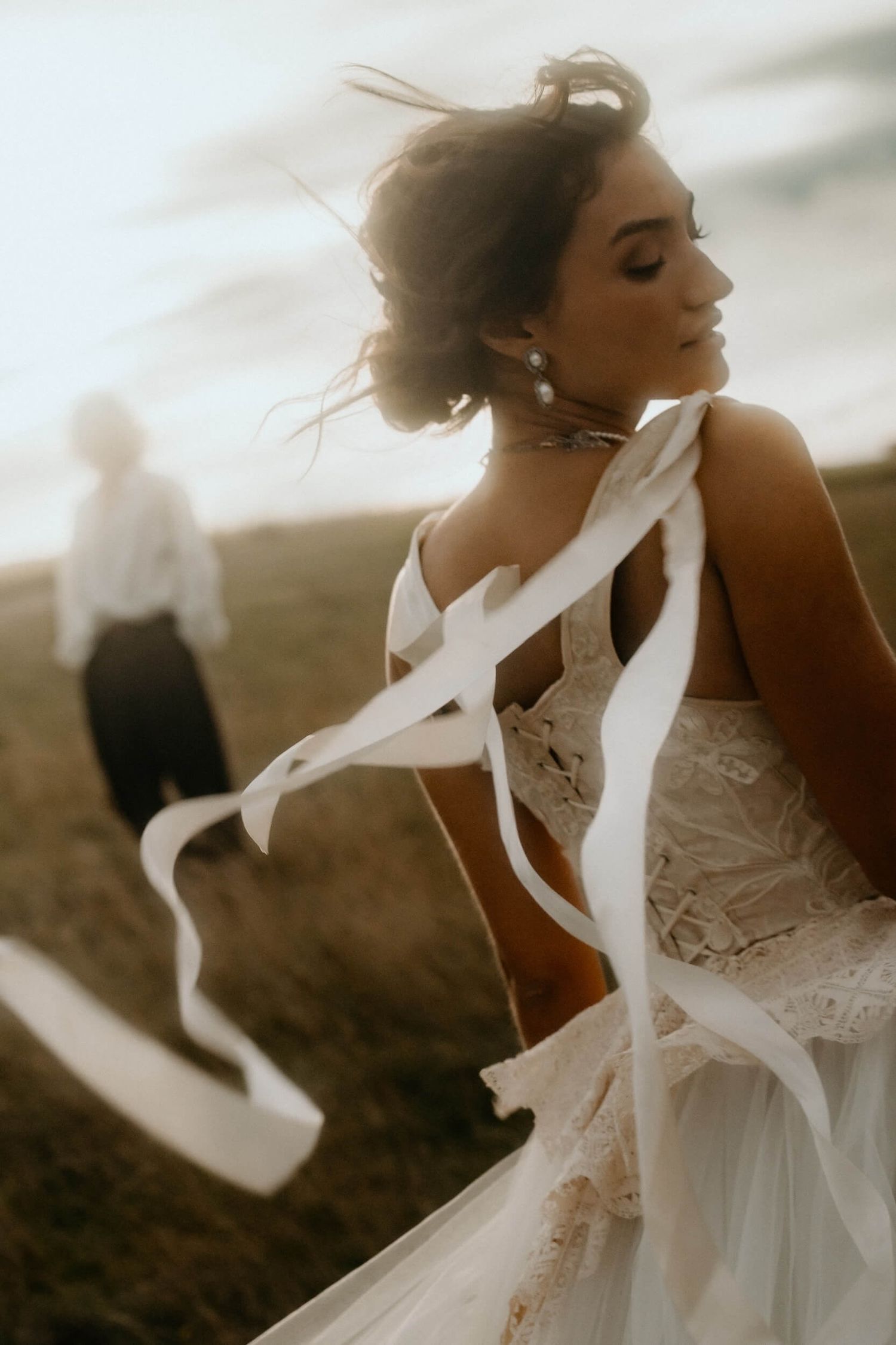 Bride in lace wedding dress with flowing ribbons blowing in wind in open field.