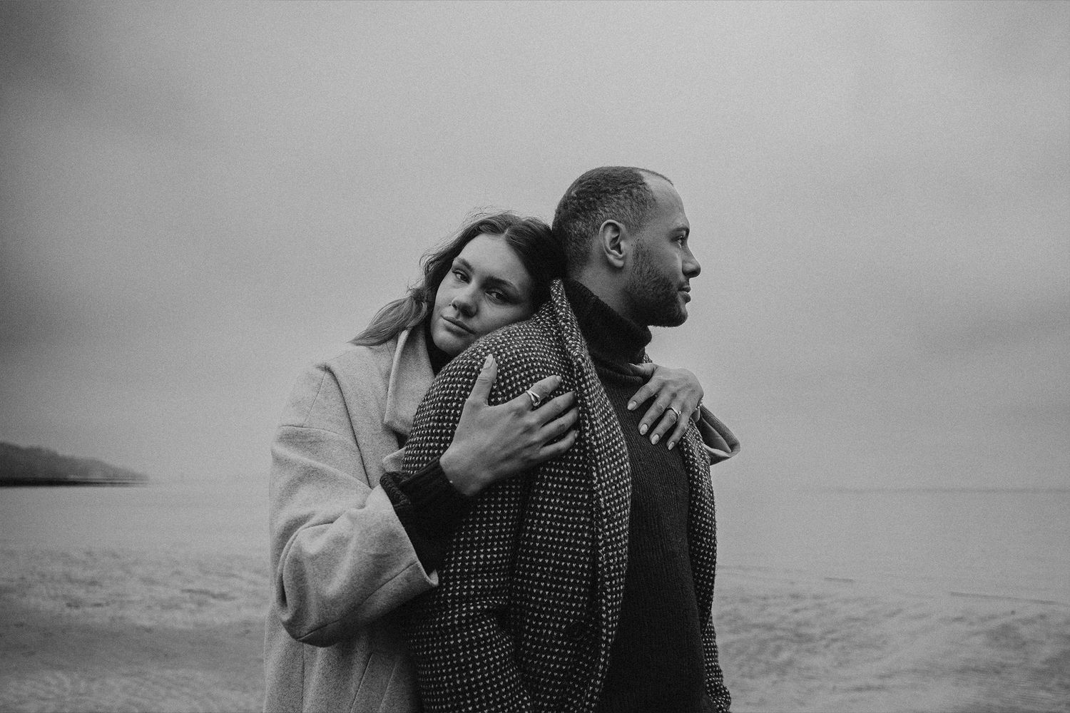 Black and white photo of a couple embracing on a cloudy beach, woman resting her head on mans shoulder.