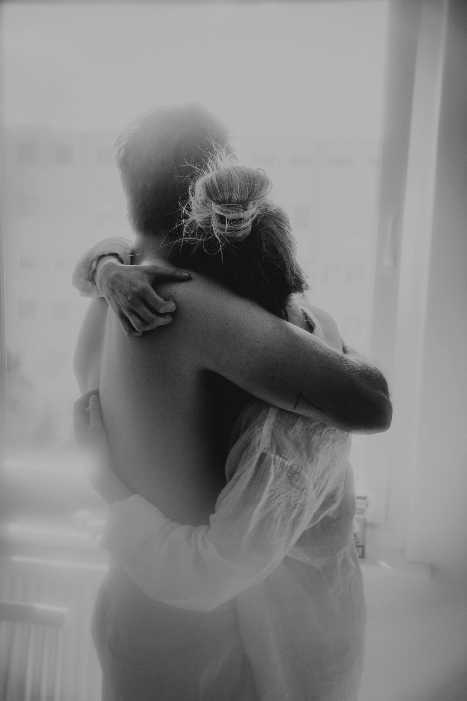 Black and white photo of two people sharing an intimate embrace near a window in soft natural light.
