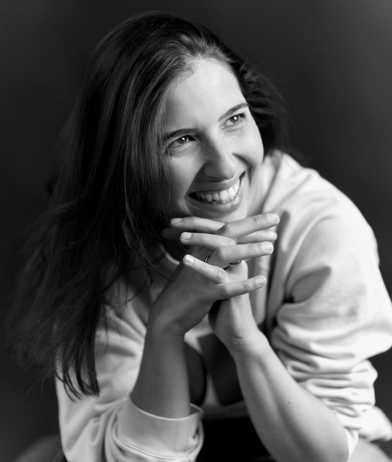 Black and white portrait of smiling young woman with hands clasped under chin.