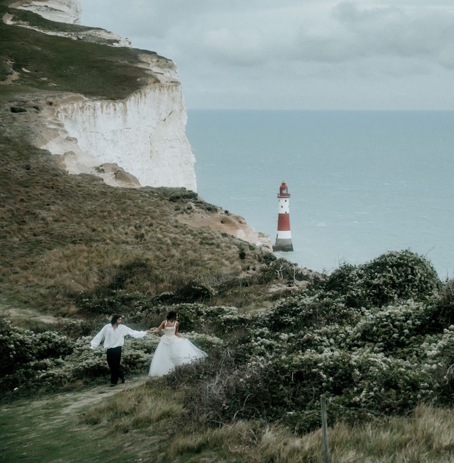 Bride and groom walk hand in hand along cliffs near Beachy Head lighthouse, Sussex, England.
