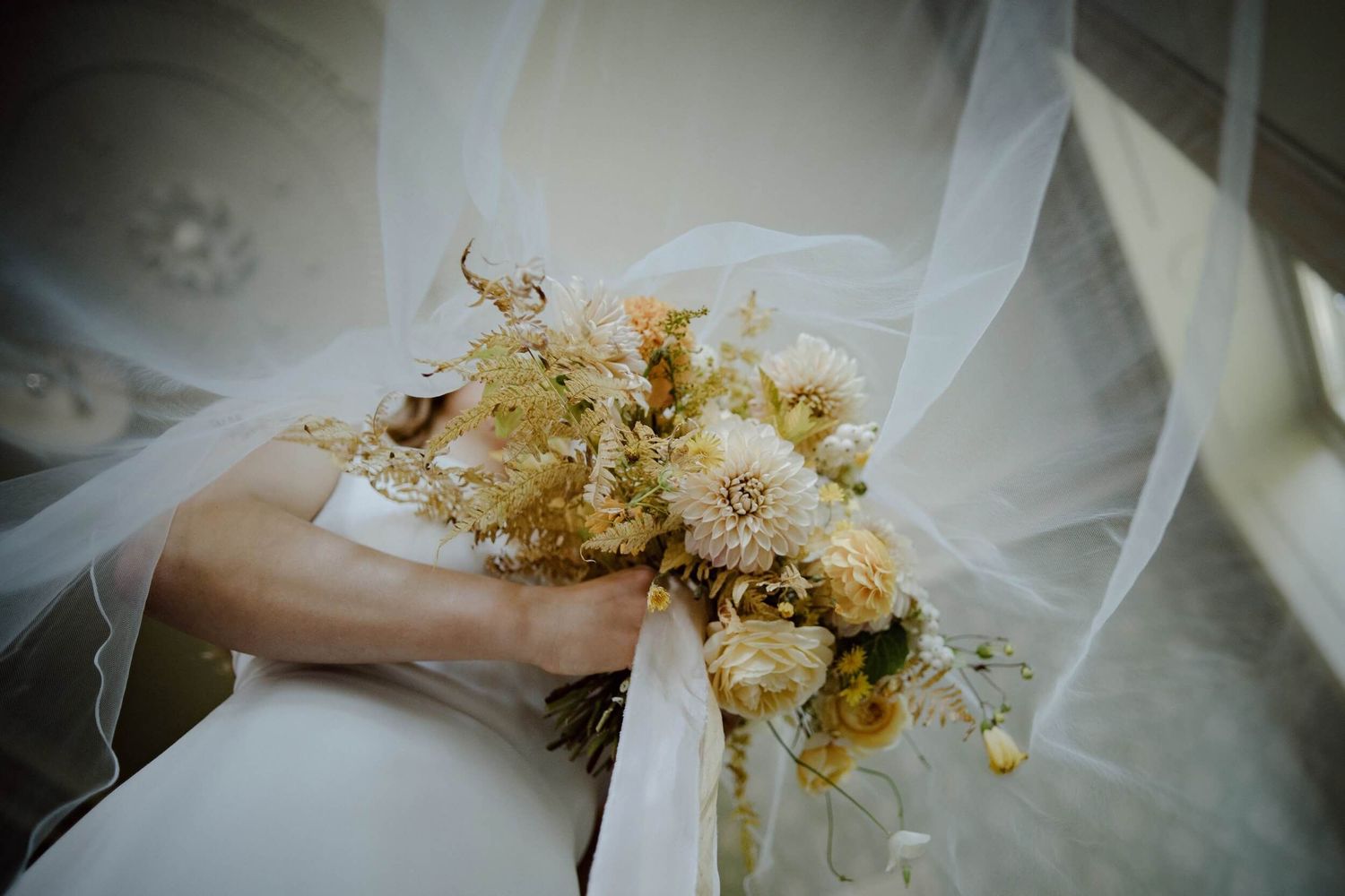 Bride holding a dried floral bouquet of cream and yellow flowers beneath a soft tulle veil.