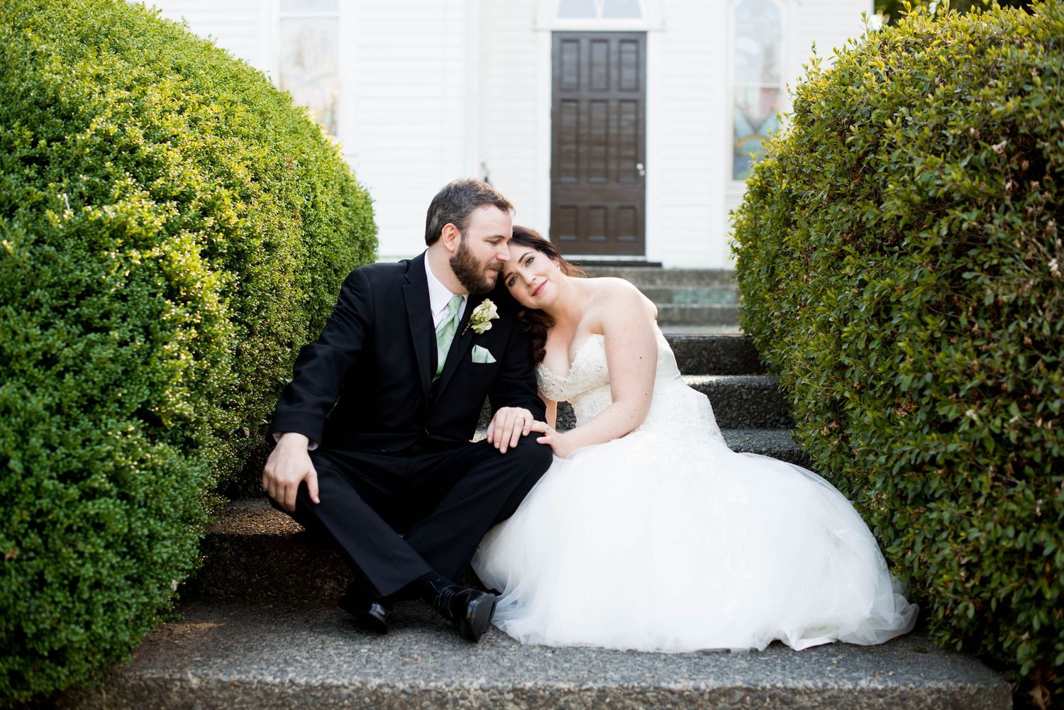 Bride and groom sitting together on stone steps surrounded by green hedges during their wedding portraits.