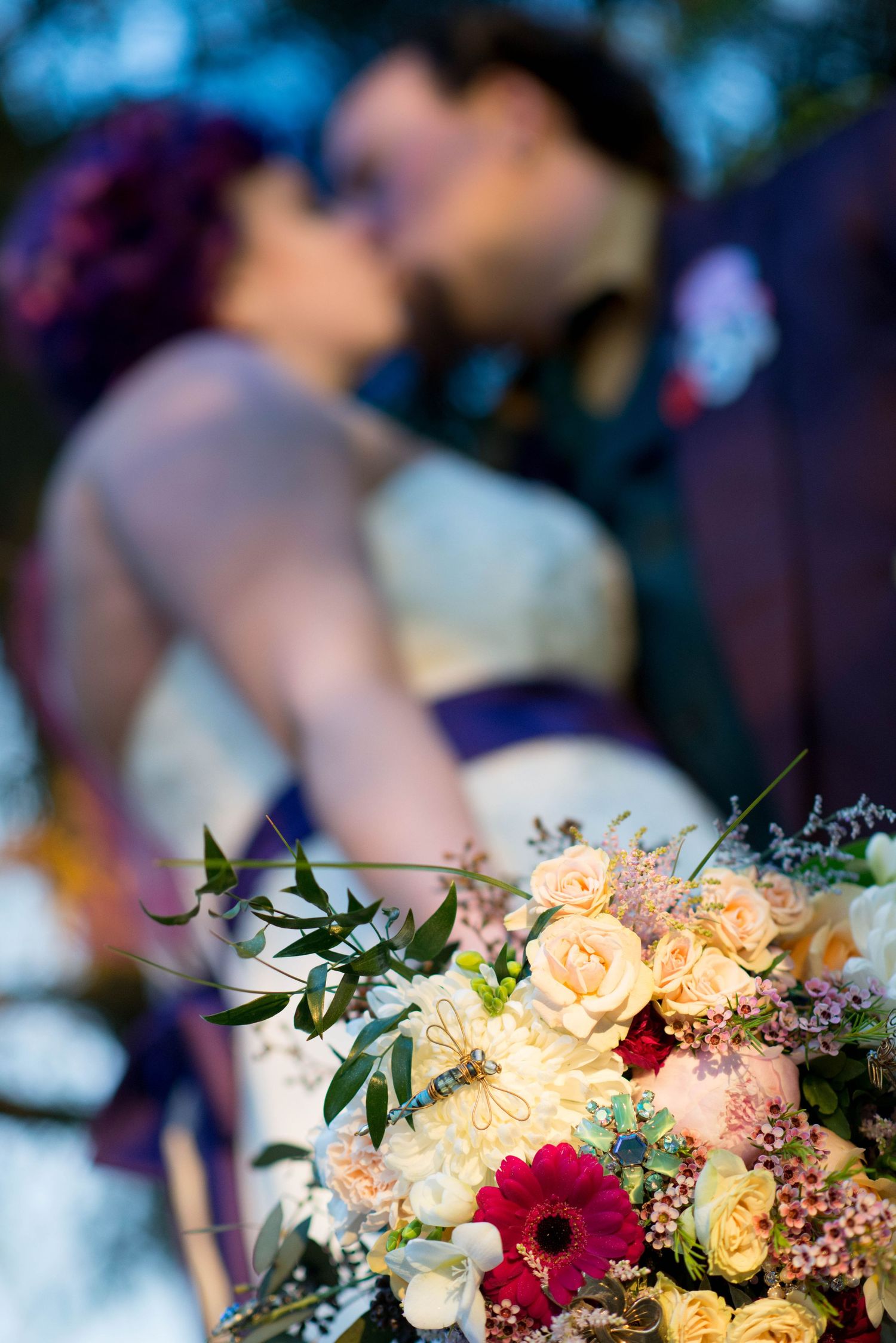 Wedding bouquet of roses and gerbera daisies with a kissing couple softly blurred behind in Abbotsford, British Columbia.