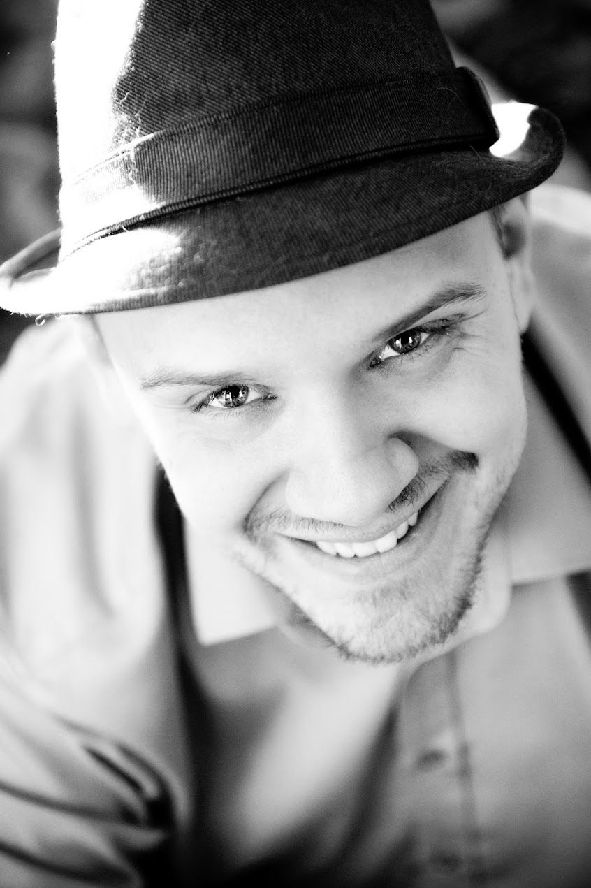 Black-and-white portrait of a smiling young man wearing a fedora outdoors.