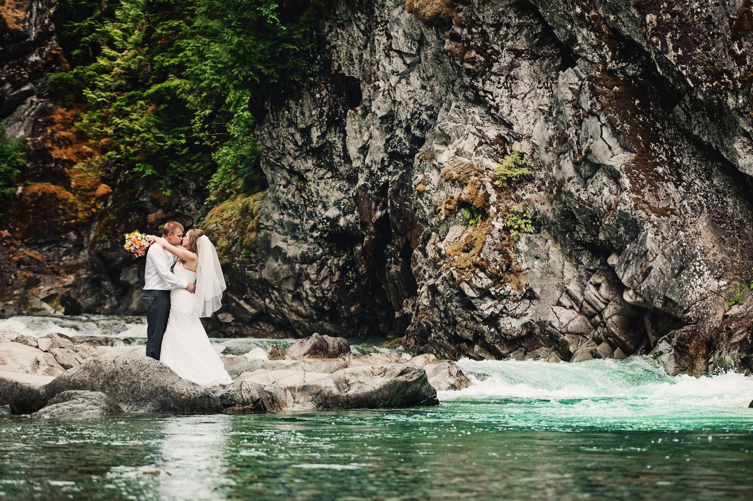 Bride and groom embracing beside a mountain river during their wedding portraits in Trail, British Columbia.