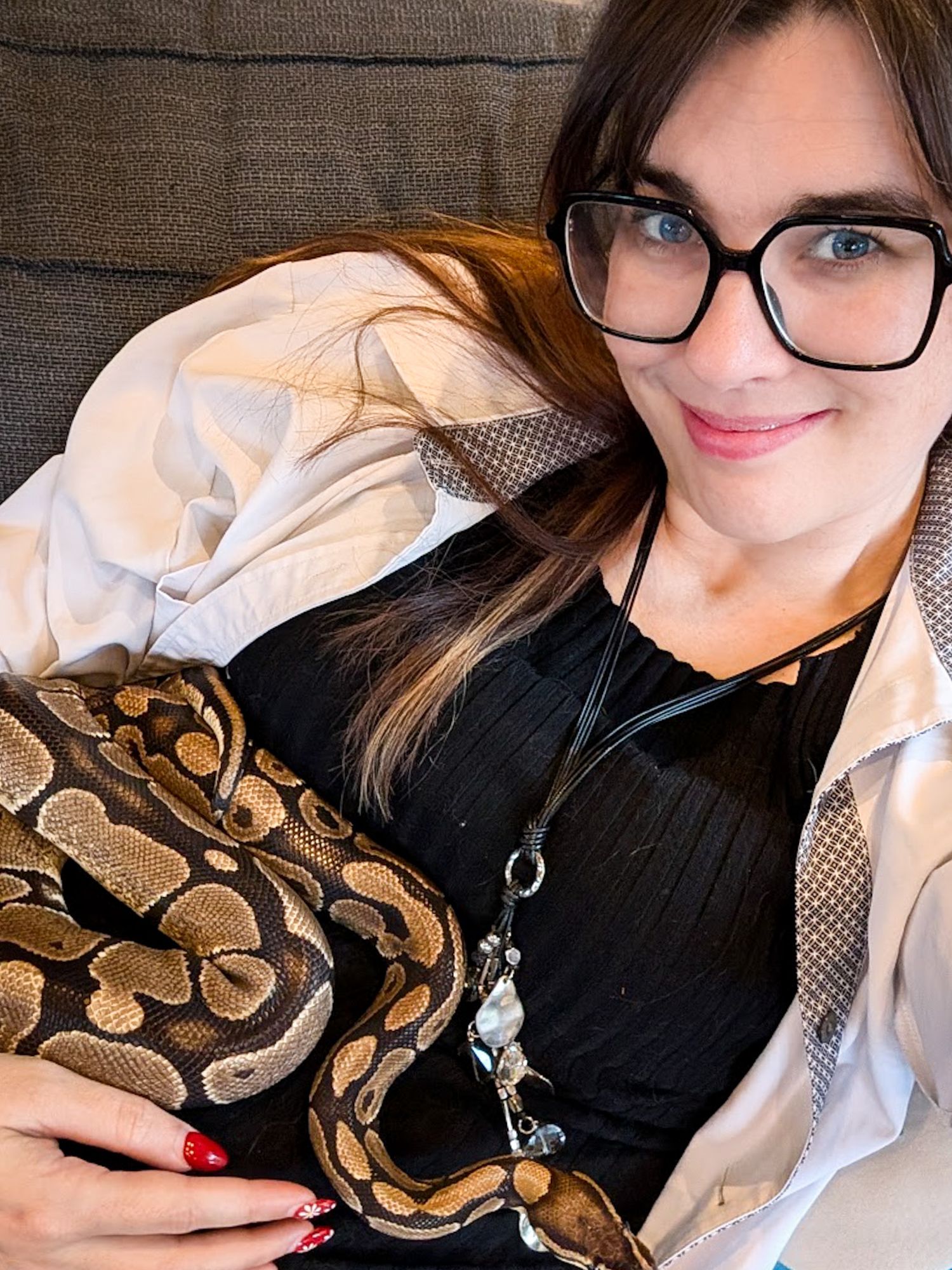 Smiling woman with black glasses cradling a ball python snake while relaxing on a sofa.