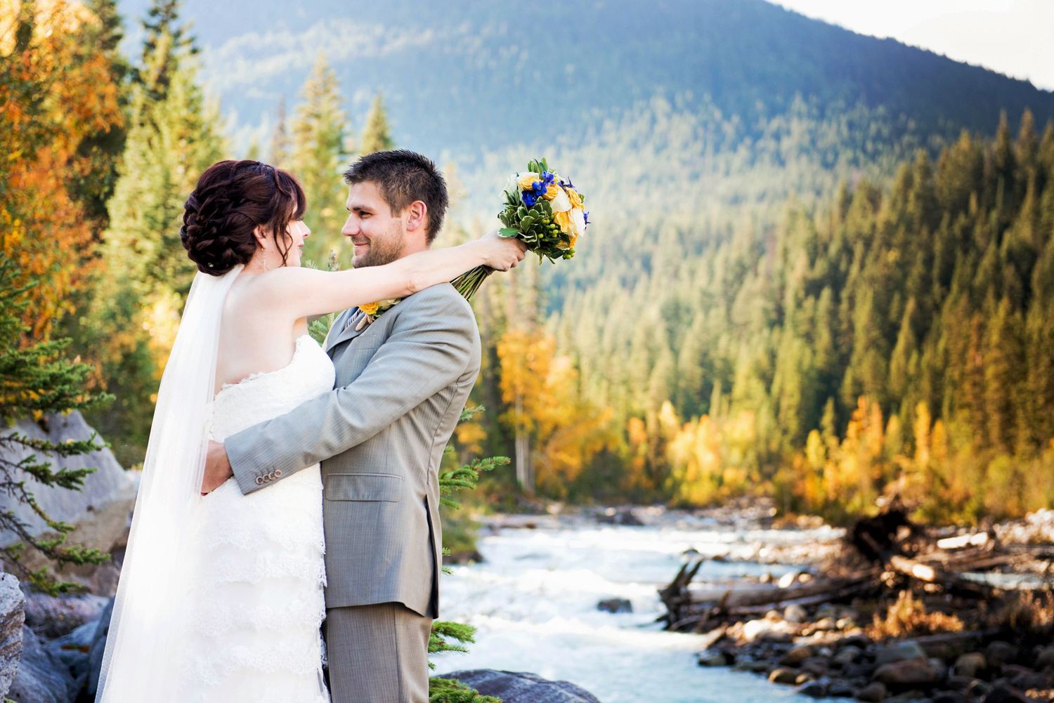 Bride and groom embracing beside a mountain river during their wedding portraits in Valemount, British Columbia.