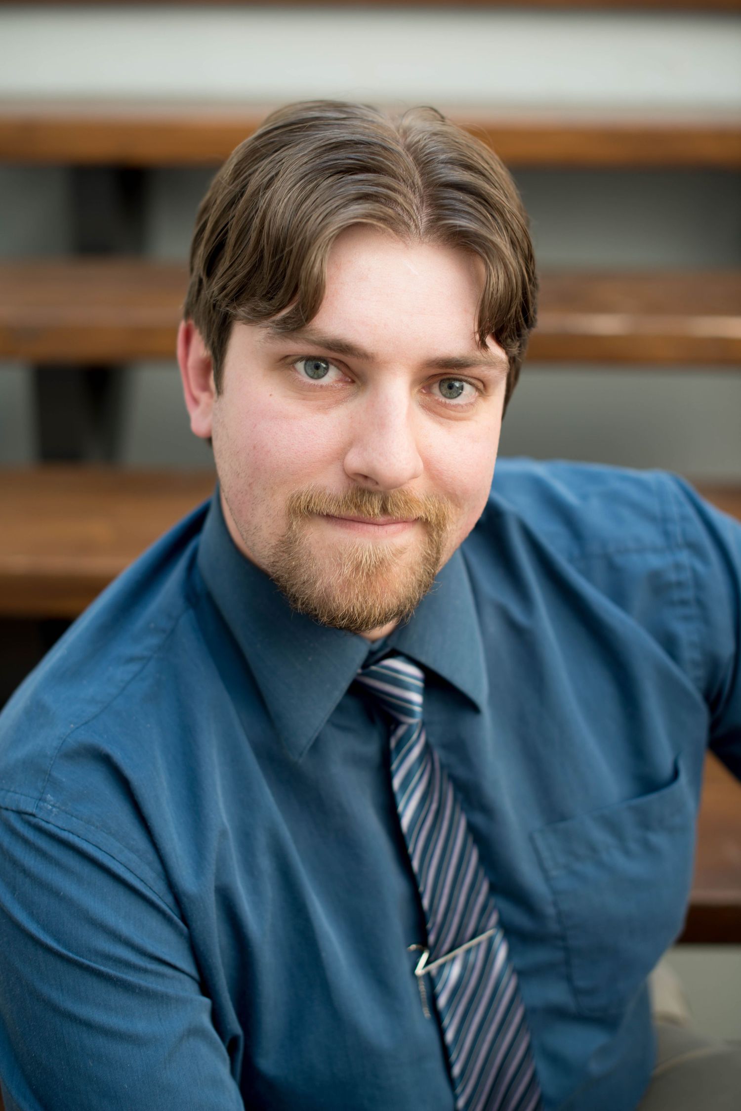 Professional headshot of bearded man in blue shirt and striped tie sitting on bleachers in Chilliwack, British Columbia.
