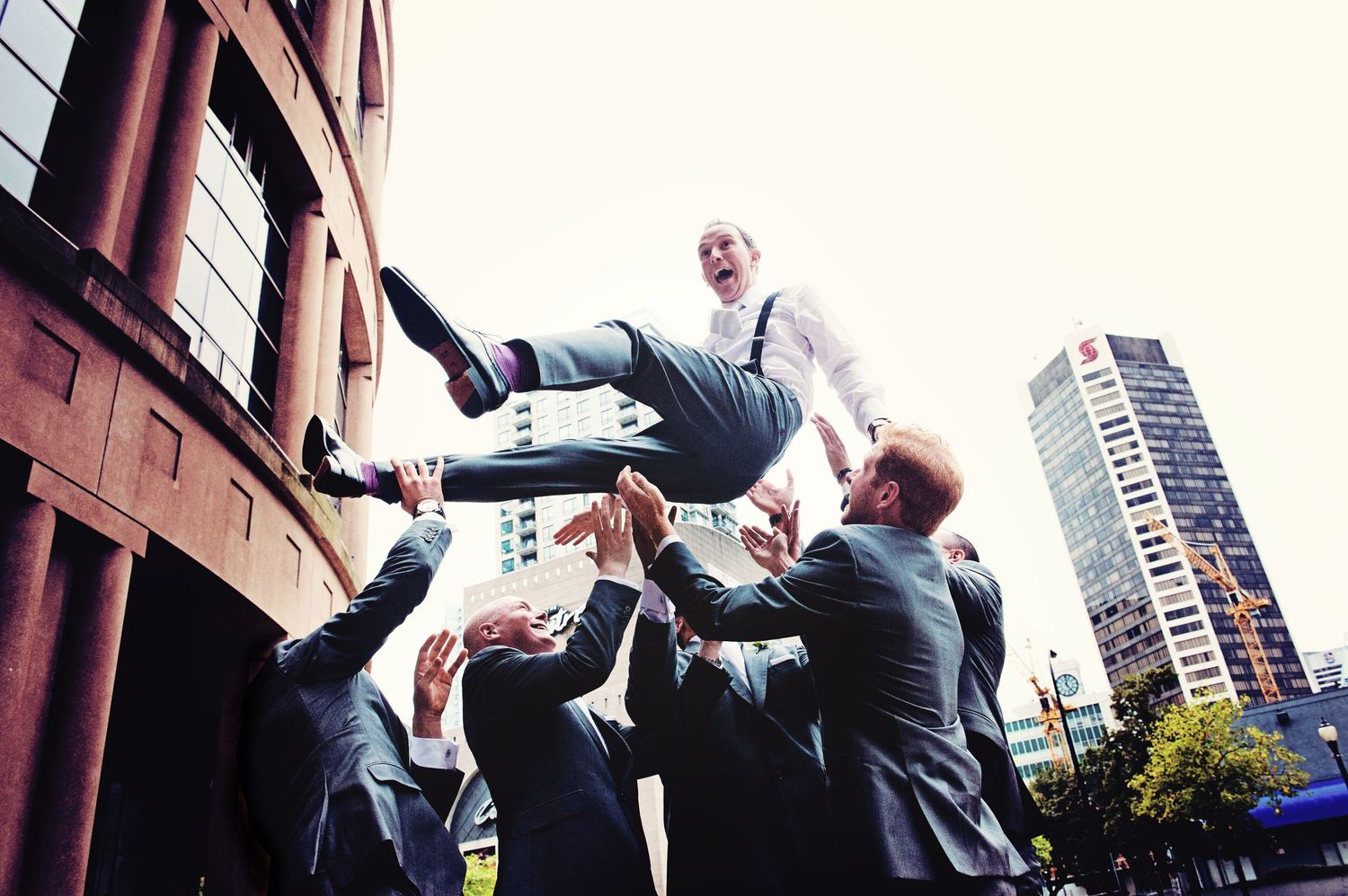 Groomsmen tossing the groom into the air outside the Vancouver Public Library during a wedding celebration.