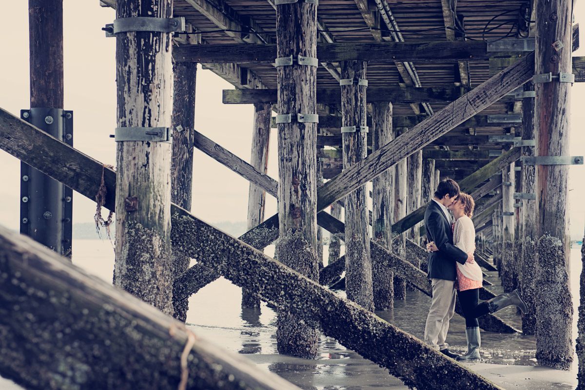 Couple sharing a kiss beneath the White Rock Pier during an engagement session in White Rock, British Columbia.