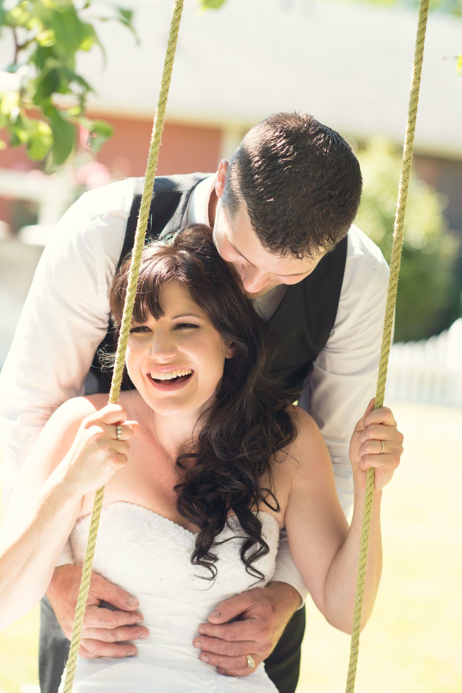 Bride laughing on a rope swing as the groom kisses her cheek during their wedding portraits.
