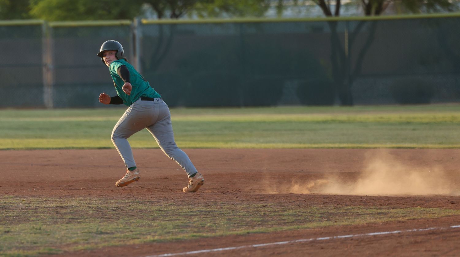 Baseball player in a teal jersey sprints toward a base as dust rises across the sunlit infield.
