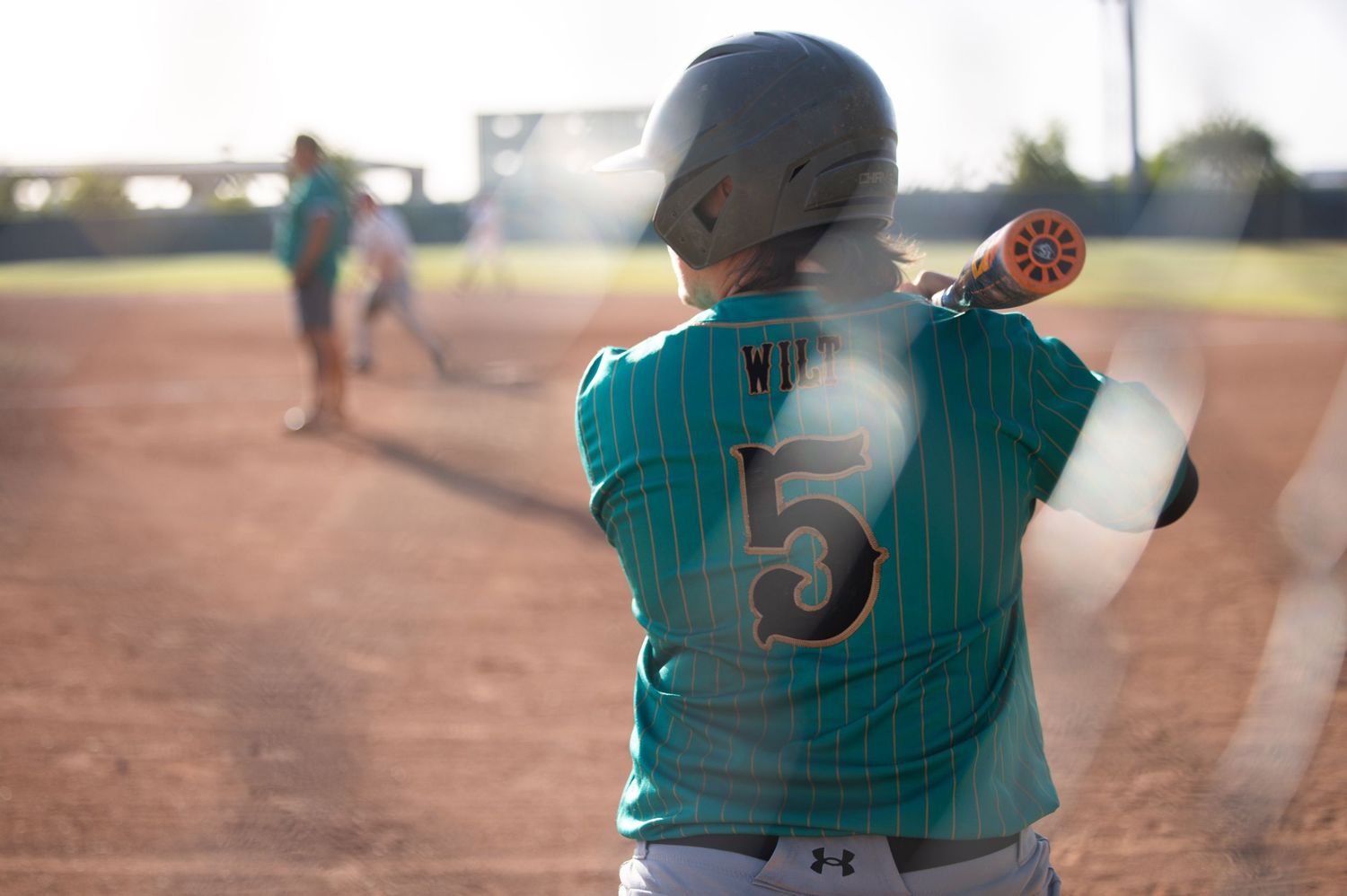 Teal-uniformed baseball player wearing number 5 prepares to throw on a sunlit diamond, viewed through a chain-link fence.