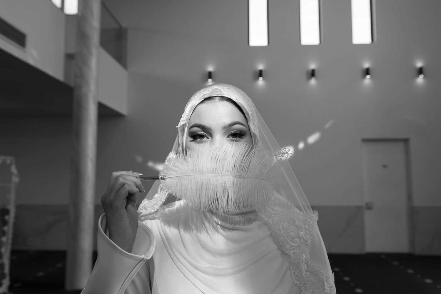Black and white photo of a woman in a white hijab holding a feather in front of her face indoors.