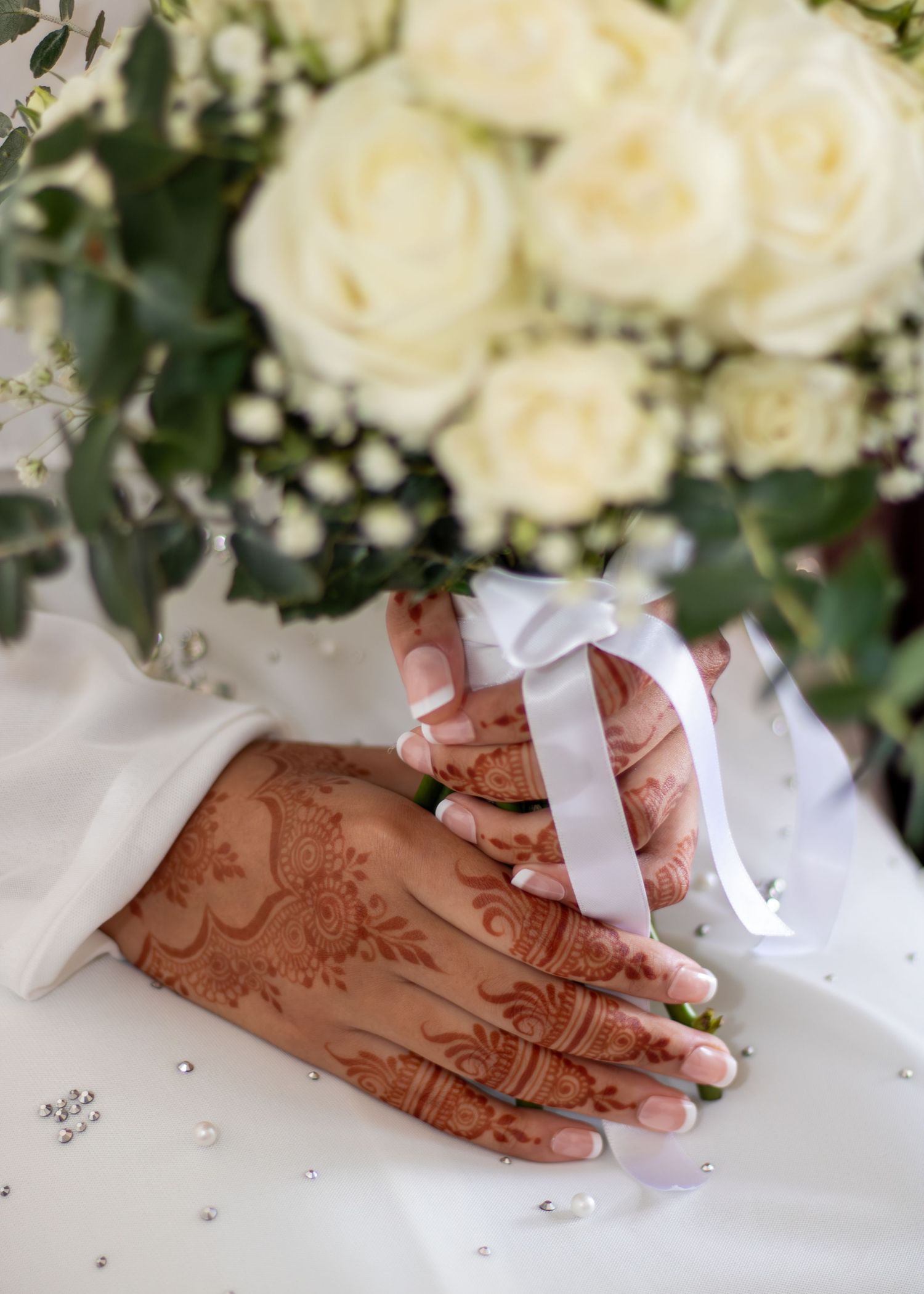 Bride's henna-adorned hands holding a white rose bouquet tied with a satin ribbon on a wedding day.