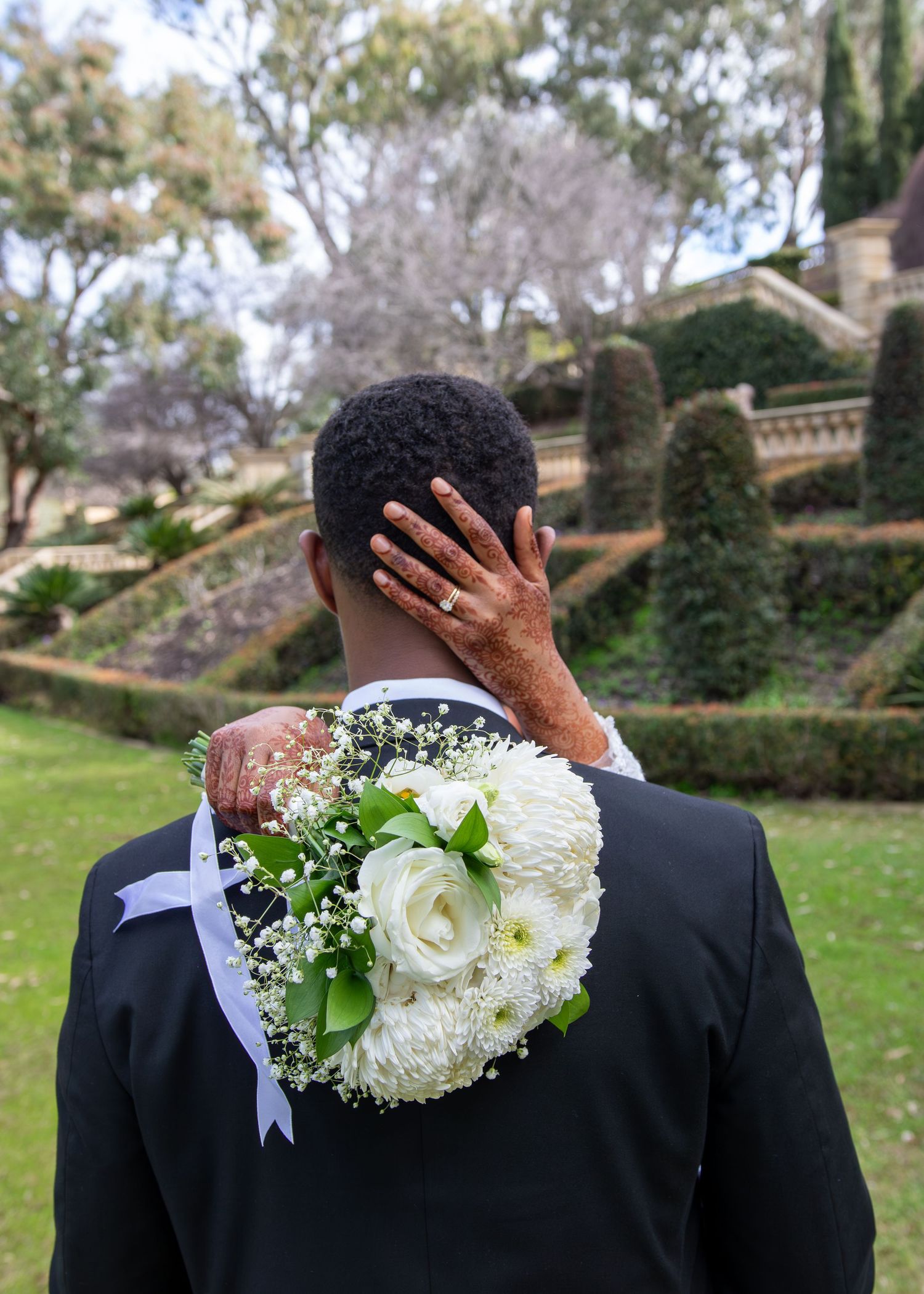 Groom in black suit viewed from behind, with bride's hand on shoulder holding white rose bouquet in garden.