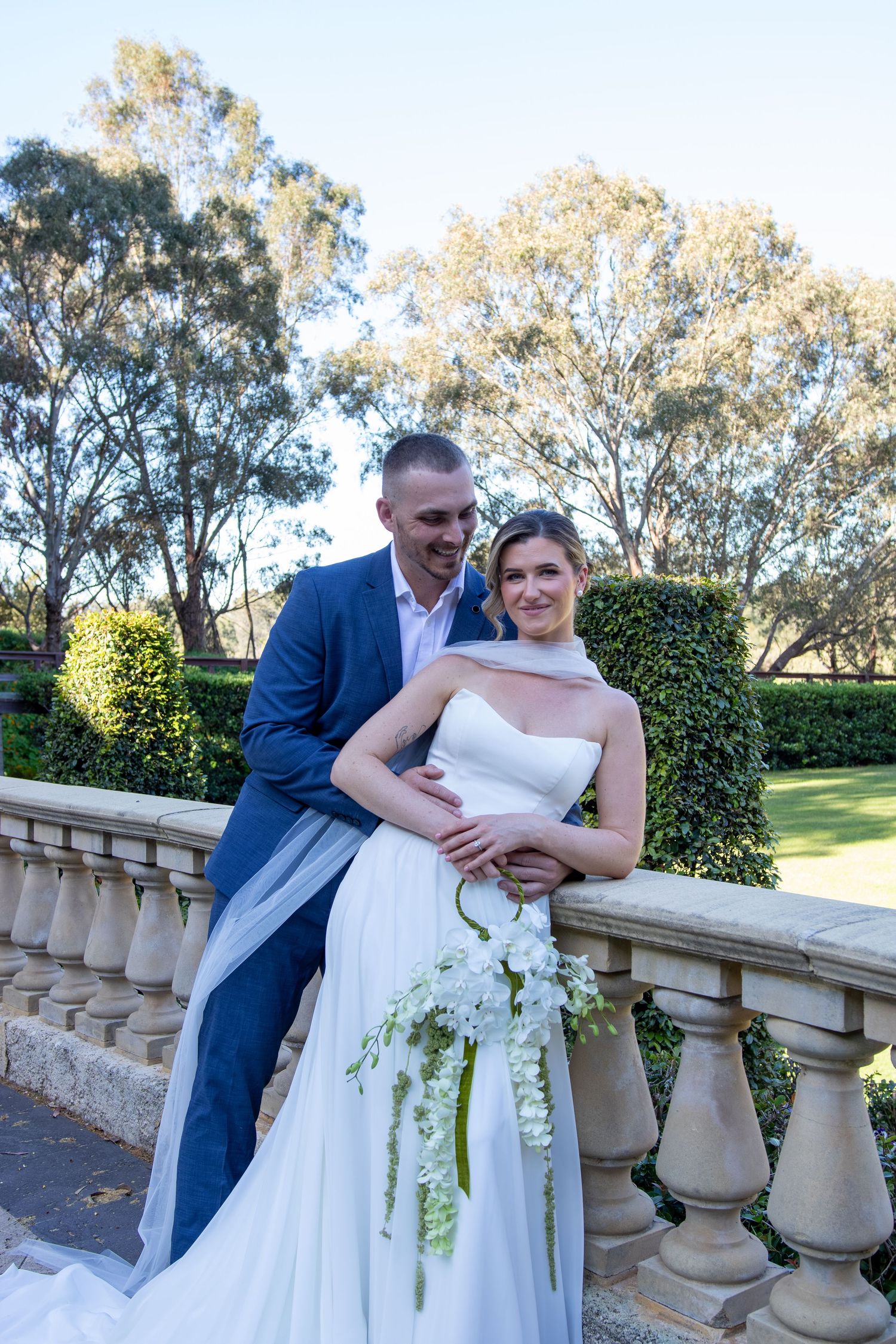 Bride and groom pose by stone balustrade in garden, she holds white bouquet, he wears blue suit.