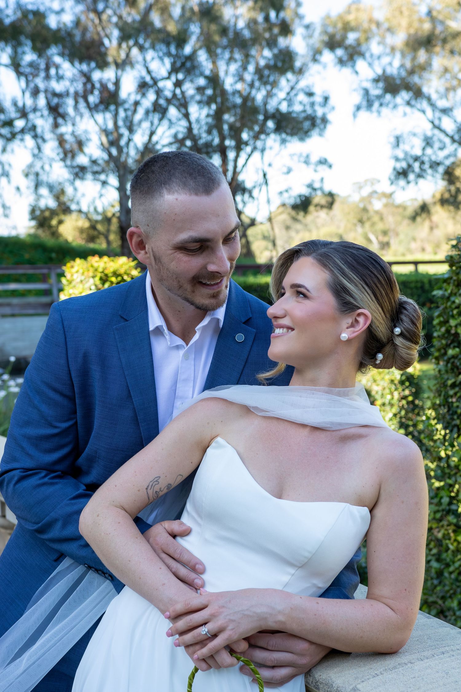 A bride and groom share a romantic moment outdoors, with the groom in a blue suit embracing the bride in a white gown.