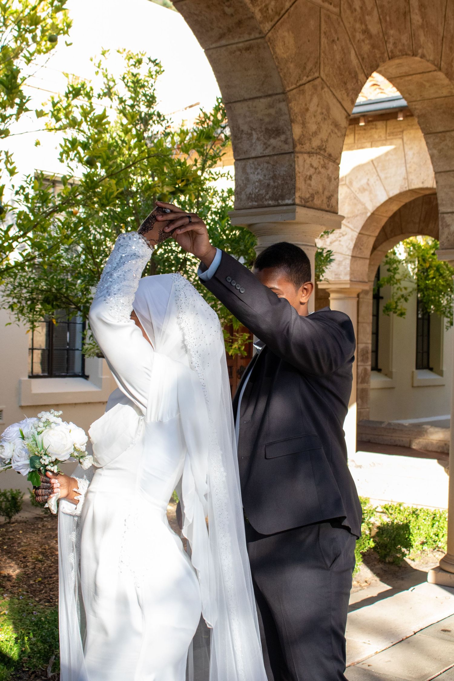 Bride and groom dancing under stone arches, bride in white gown with veil holding bouquet, groom in dark suit.