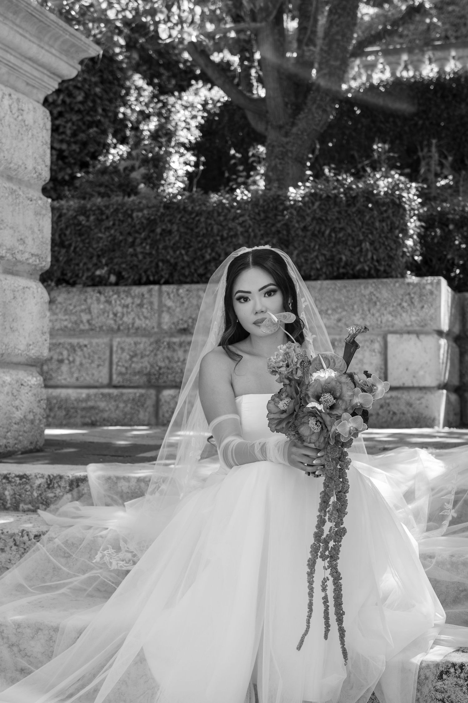 Black and white bridal portrait of Asian bride in white gown, veil, gloves, holding floral bouquet outdoors.